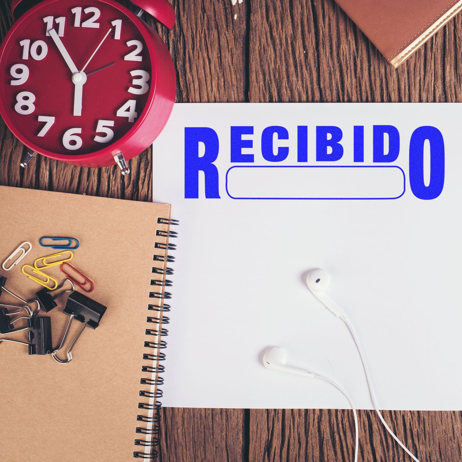 Large Recibido Rubber Stamp in blue ink on paper, surrounded by a red clock, notebook, paper clips, binder clips, and earphones.