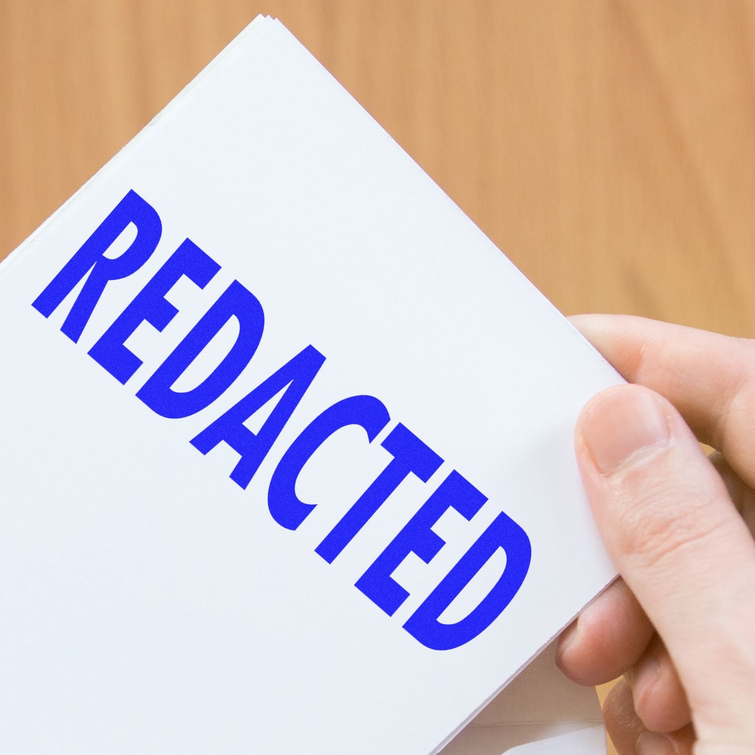 Hand holding a document stamped with REDACTED in bold blue letters using the Large Redacted Rubber Stamp against a wooden background.