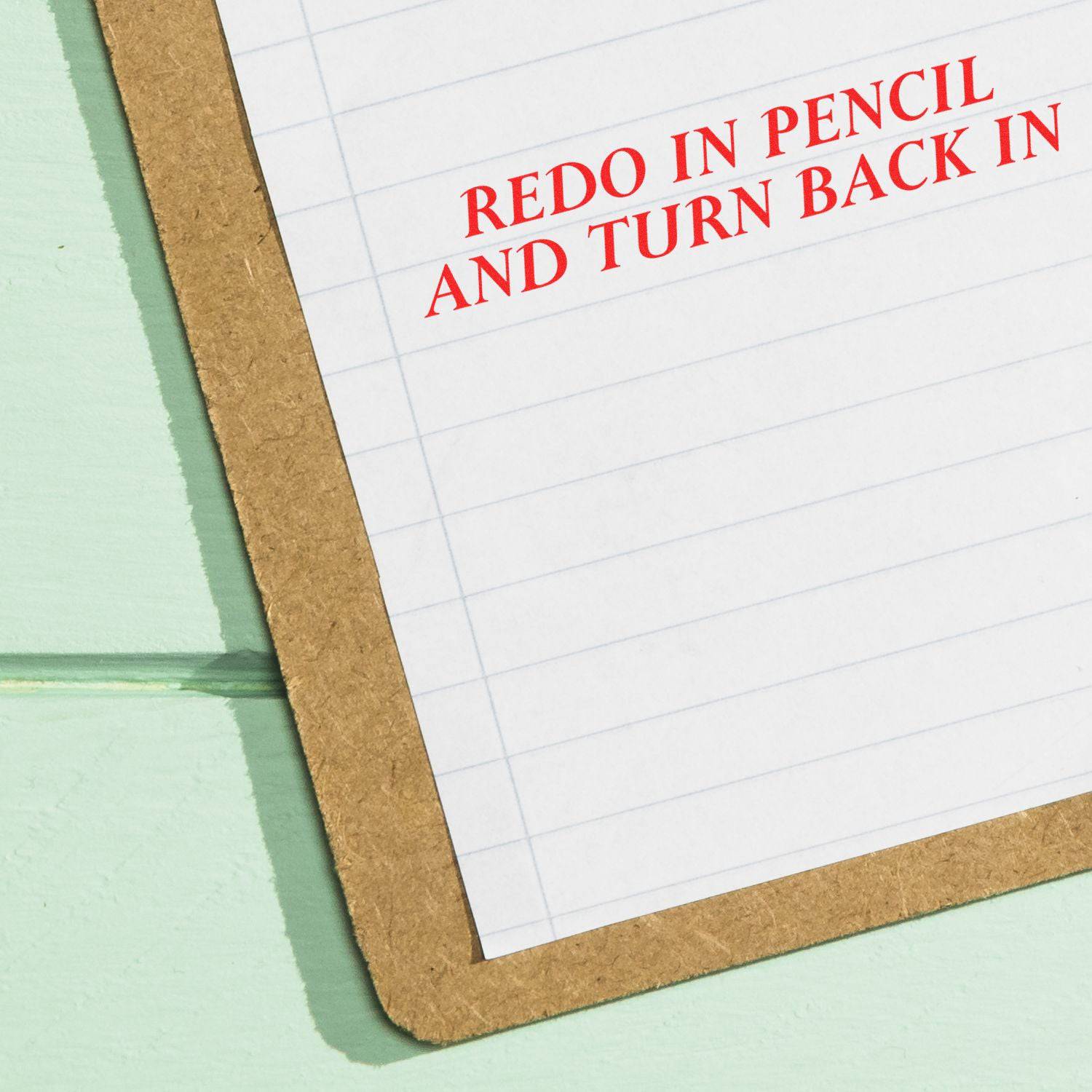 Large Redo In Pencil And Turn Back In Rubber Stamp in red ink on a lined paper clipped to a brown clipboard on a light green surface.