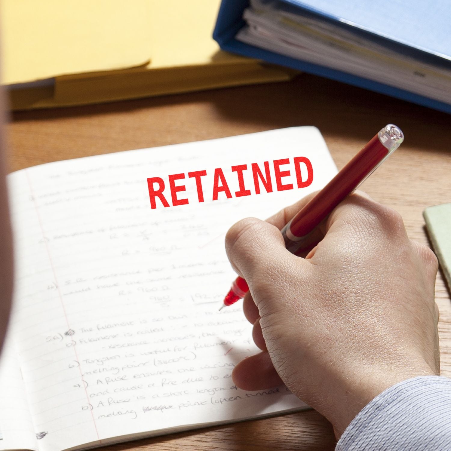 Hand holding a red pen next to a notebook stamped with RETAINED using the Large Retained Rubber Stamp, with folders in the background.