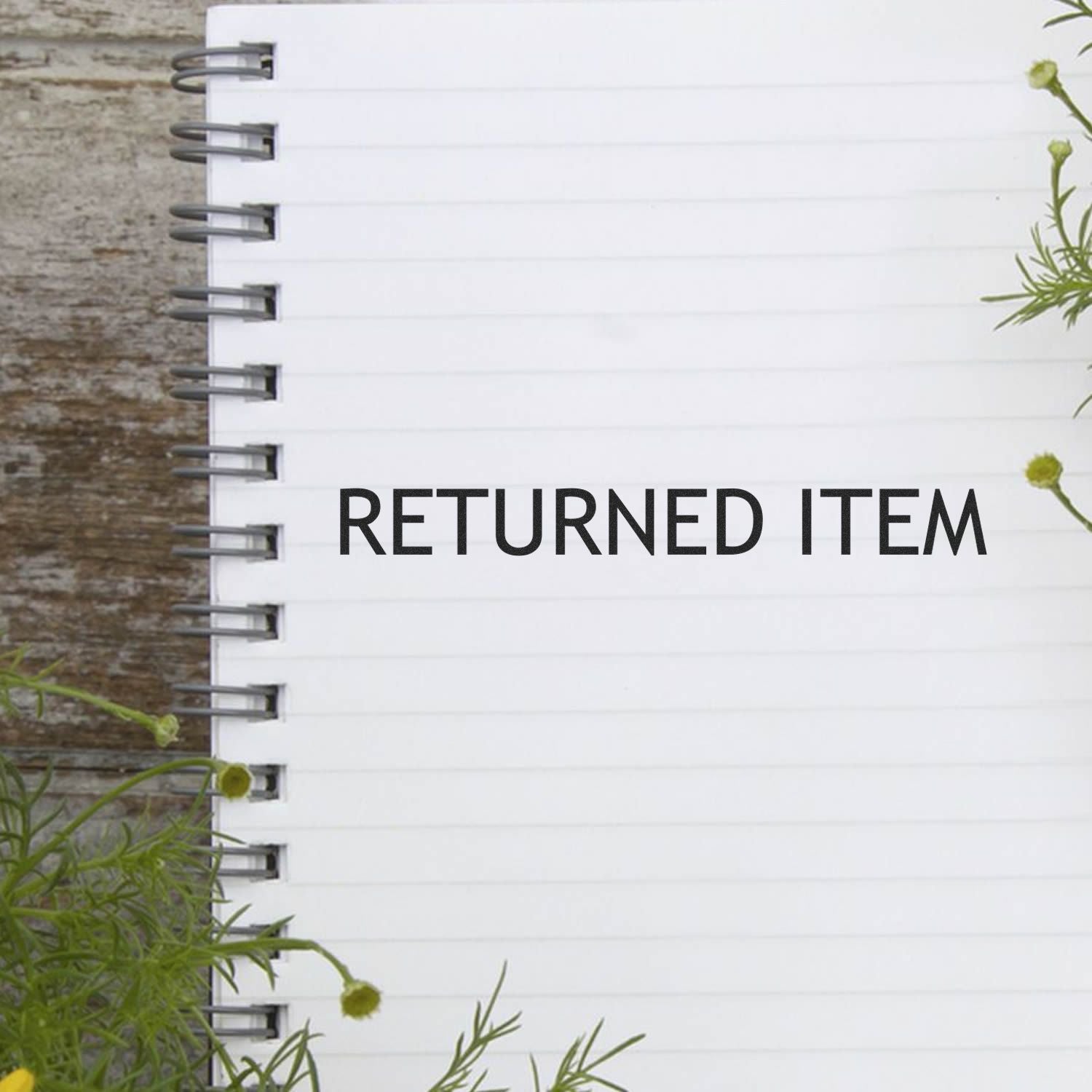 Spiral notebook with 'RETURNED ITEM' stamped in black ink, surrounded by green plants on a wooden surface.