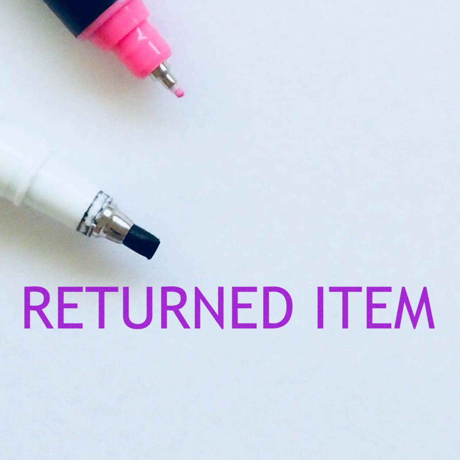 Large Returned Item Rubber Stamp in use, displaying the text RETURNED ITEM in purple ink on white paper, with two markers nearby.