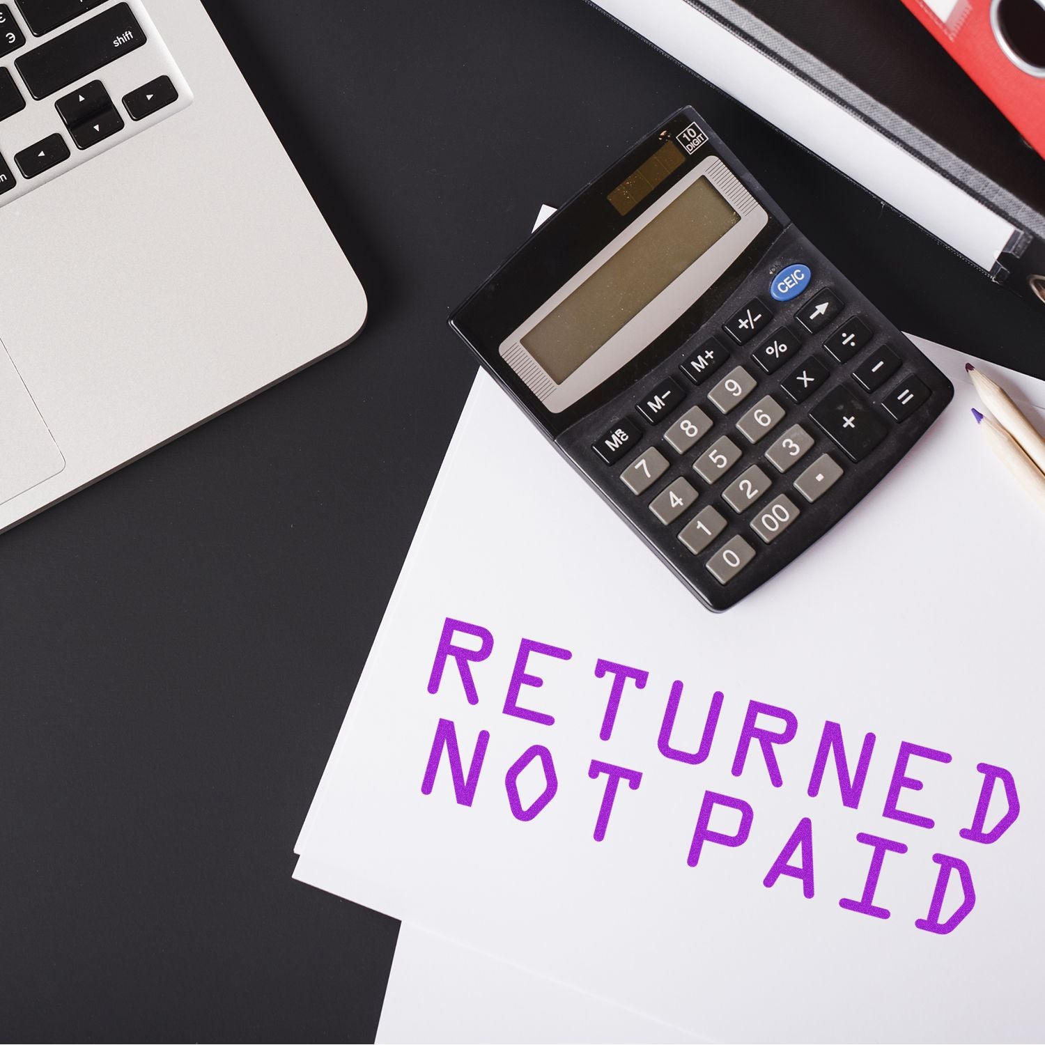 Slim Pre-Inked Returned Not Paid Stamp on white paper next to a calculator and laptop on a black desk.