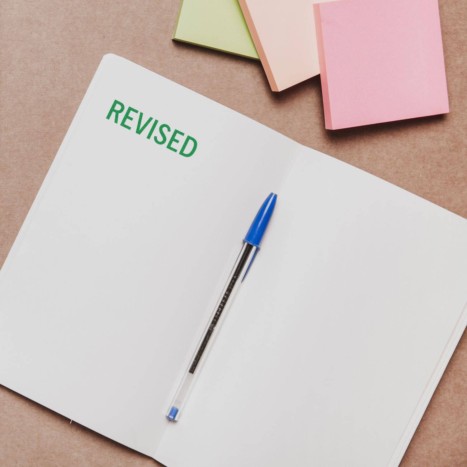 Large Revised Rubber Stamp marking REVISED in green on an open notebook, with a blue pen and colorful sticky notes nearby.