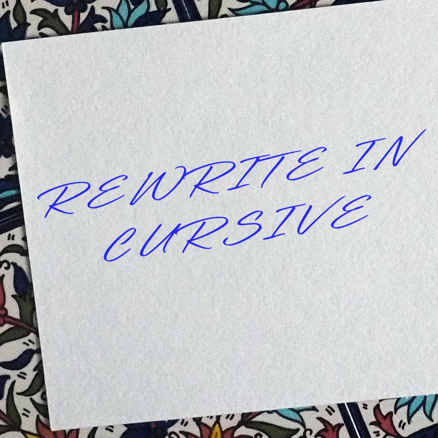 A white paper with REWRITE IN CURSIVE written in blue ink using the Large Self Inking Rewrite In Cursive Stamp, placed on a floral background.