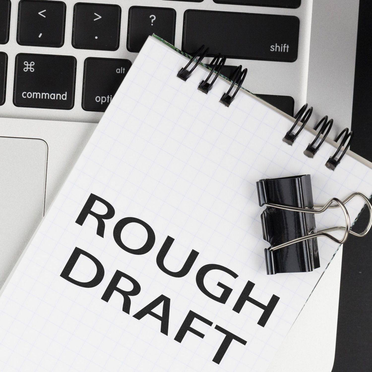 A Large Rough Draft Rubber Stamp imprint on a notepad with a binder clip, placed on a laptop keyboard.