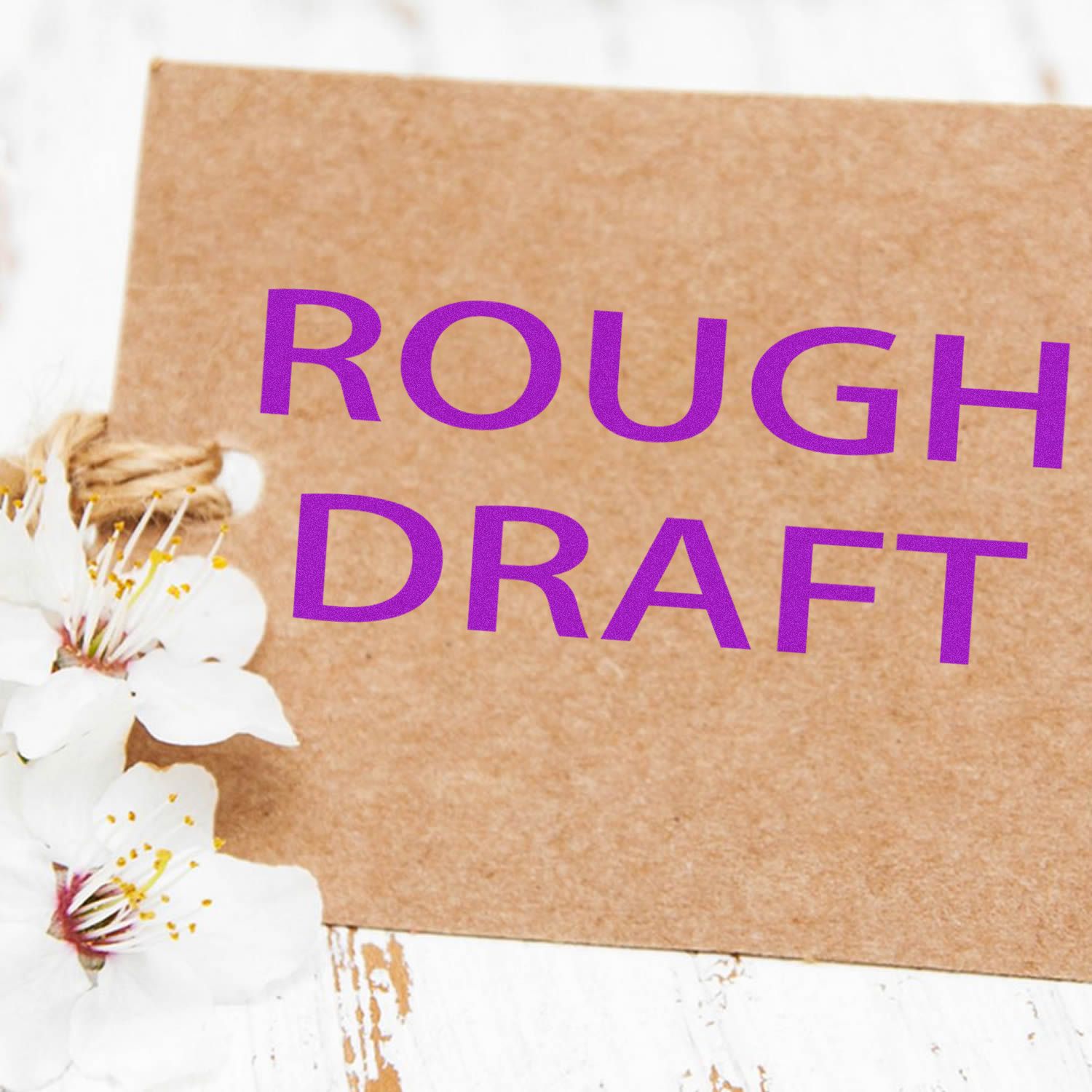 Brown card with purple 'Rough Draft' rubber stamp text, placed on a white wooden surface with white flowers.