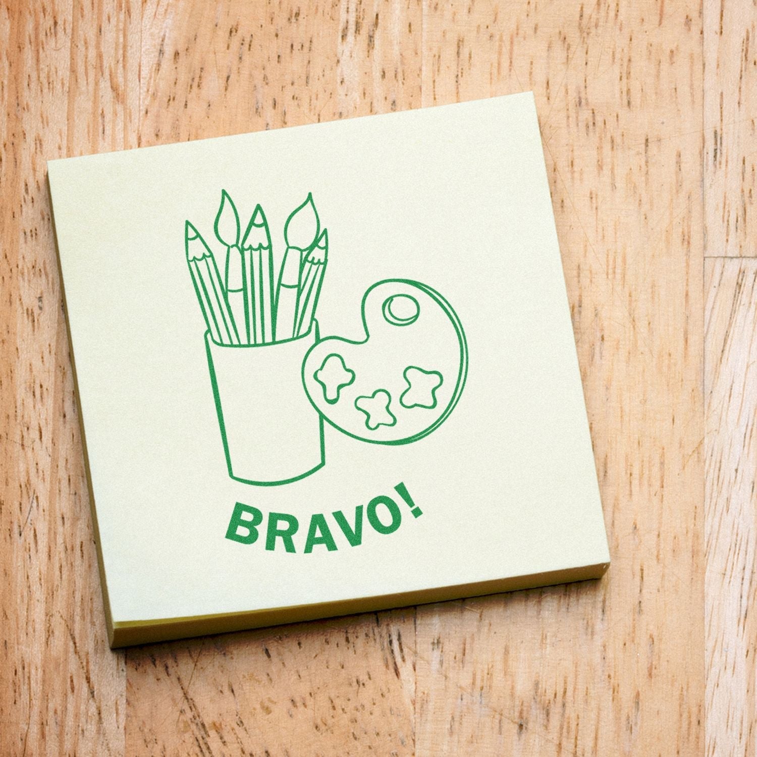 A green stamped image of art supplies and the word BRAVO! on a sticky note, created with the Self Inking Round Bravo Stamp, on a wooden surface.