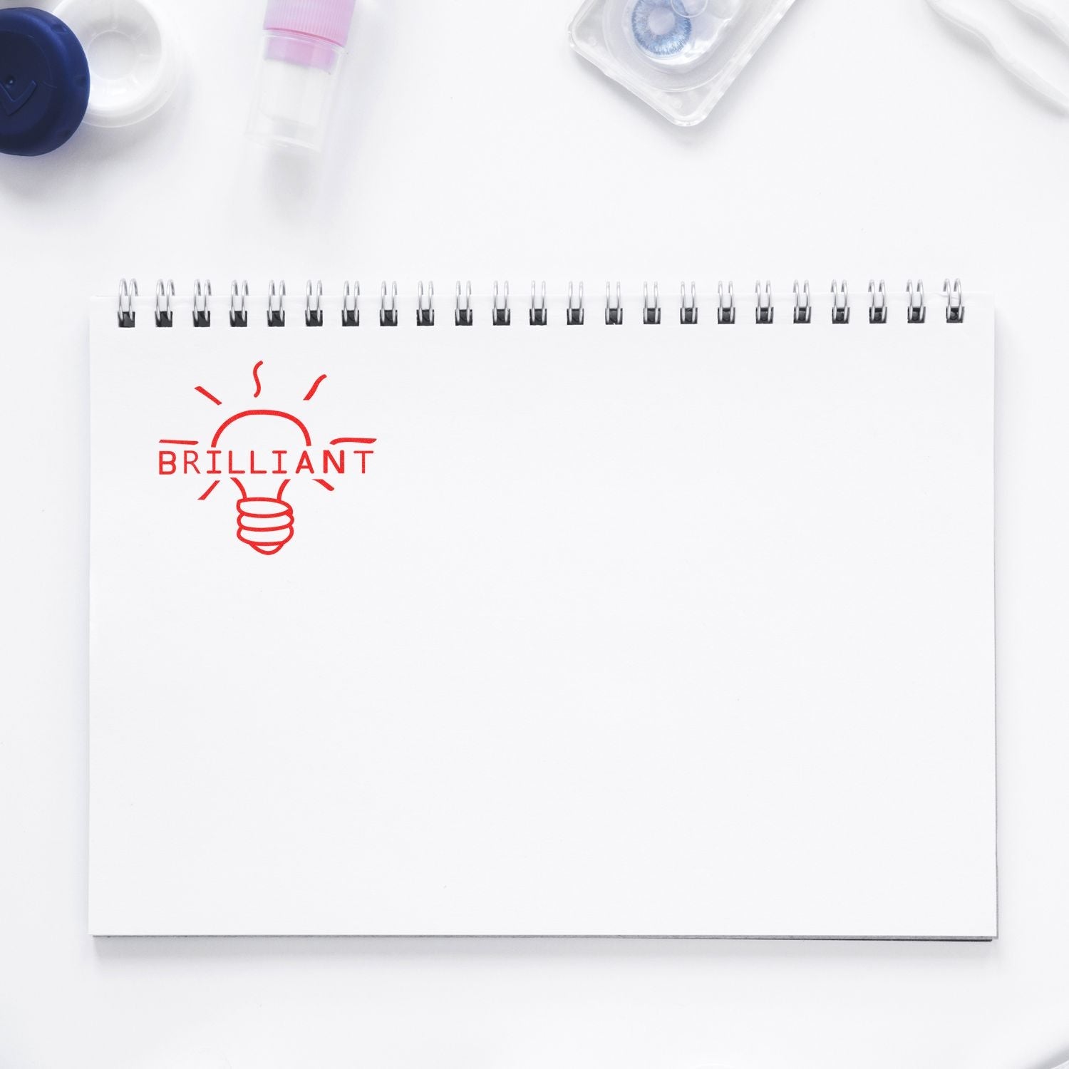 Self Inking Round Brilliant Stamp imprinting a red BRILLIANT lightbulb design on a white spiral notebook, surrounded by office supplies.