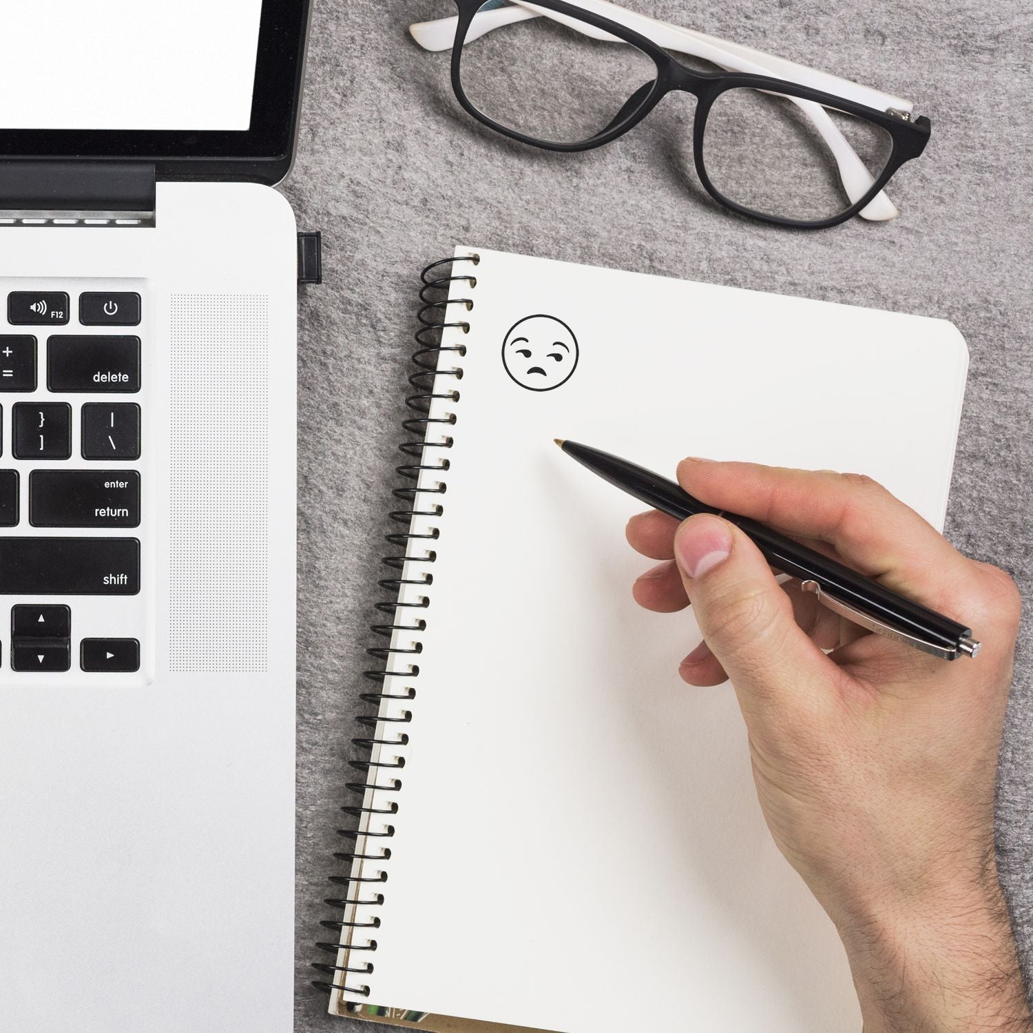 A hand using the Self Inking Round Calm Smiley Stamp on a notebook, next to a laptop and glasses on a gray surface.