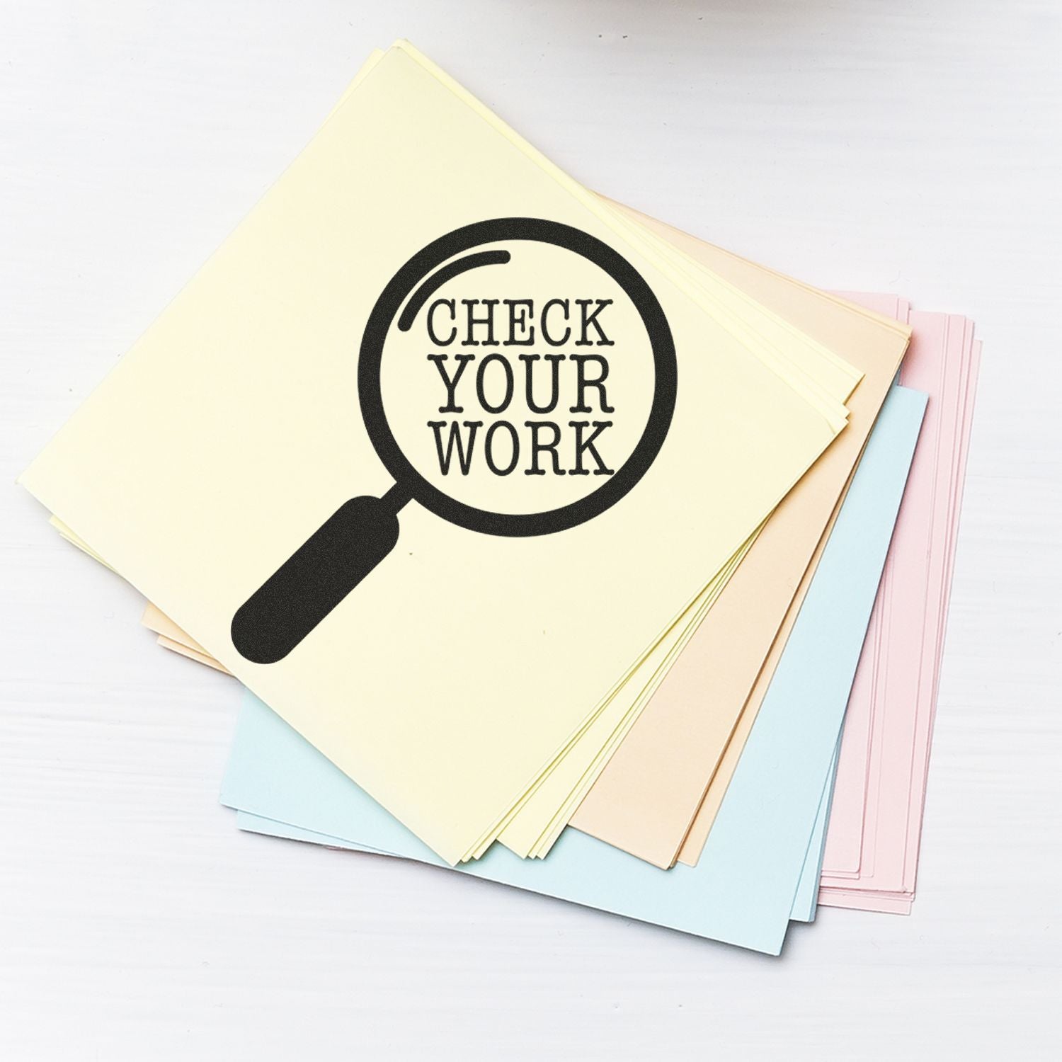 Round Check Your Work rubber stamp on a stack of pastel-colored papers, featuring a magnifying glass design.