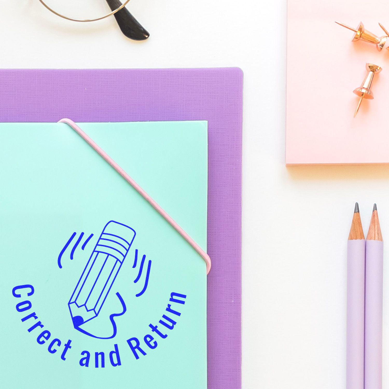 Round Correct and Return rubber stamp on a mint green folder with a purple folder underneath, surrounded by pencils and push pins.