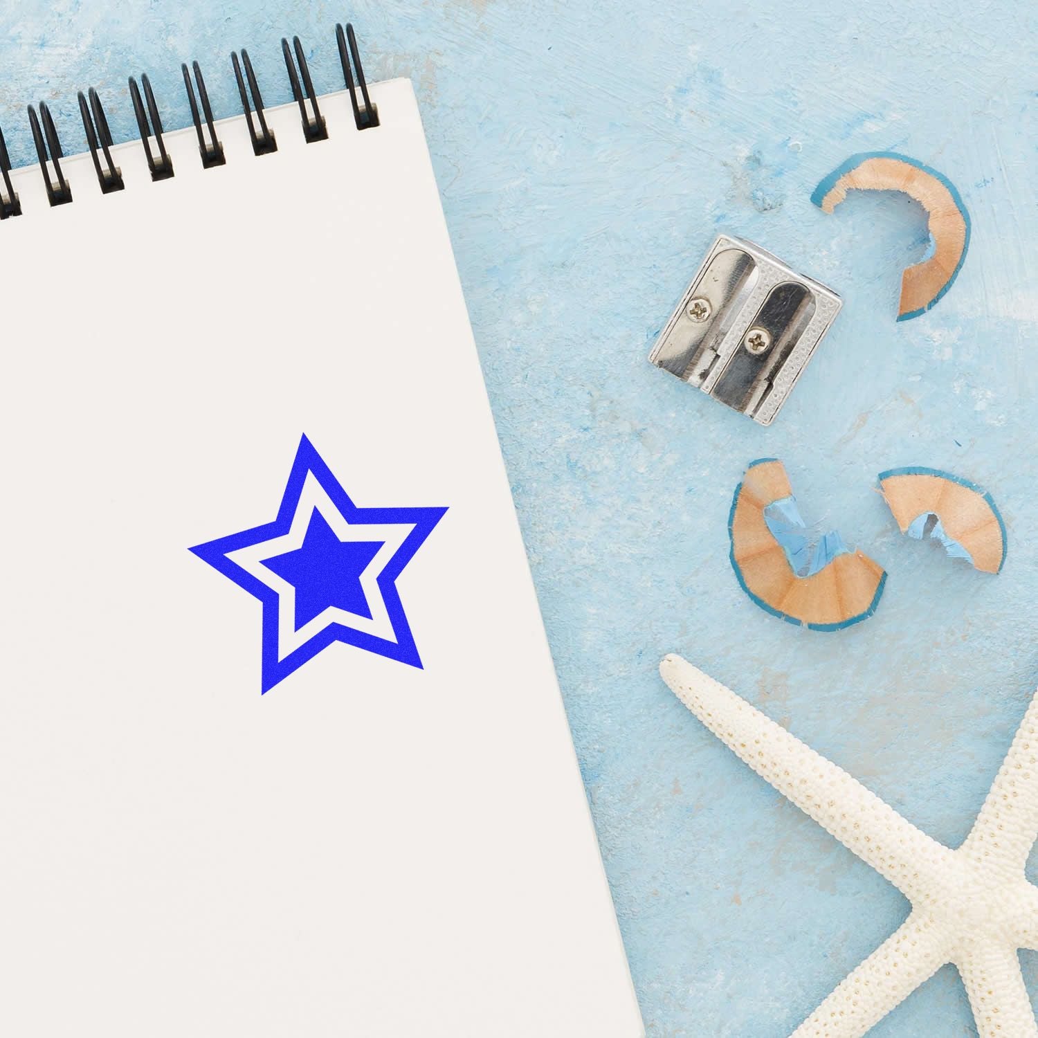 Round Double Star Rubber Stamp in blue ink on a notebook page, with a pencil sharpener and starfish nearby on a light blue surface.