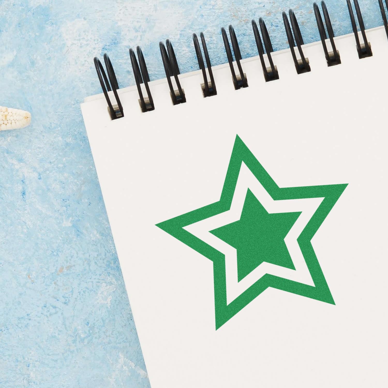 Round Double Star Rubber Stamp in green ink on a spiral-bound notebook, placed on a light blue textured surface.