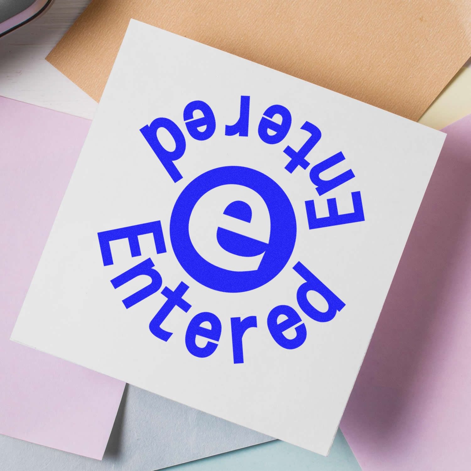 Self Inking Round Entered Entered Stamp in blue ink on a white paper, surrounded by pastel-colored papers.