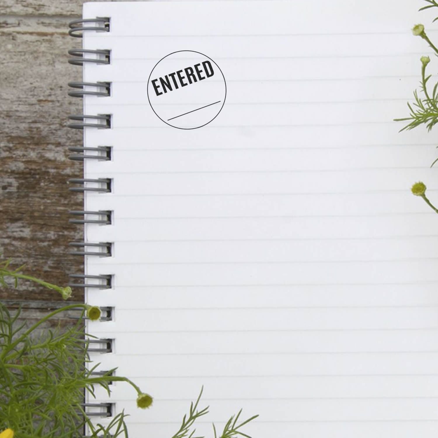 Round Entered rubber stamp mark on a blank spiral notebook page with plants in the background.