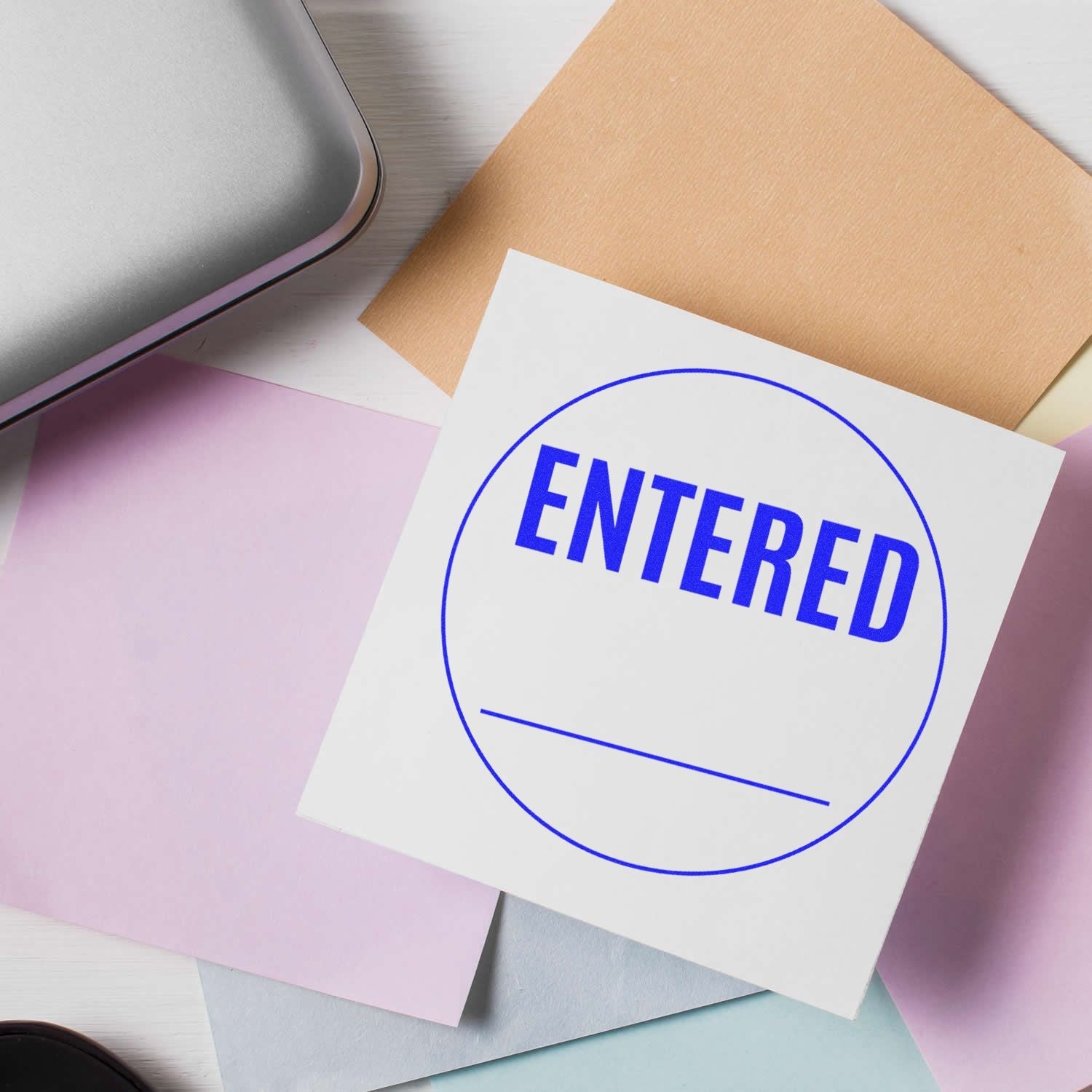 Self Inking Round Entered Stamp in blue ink on white paper, surrounded by colorful sheets and a silver laptop on a white desk.