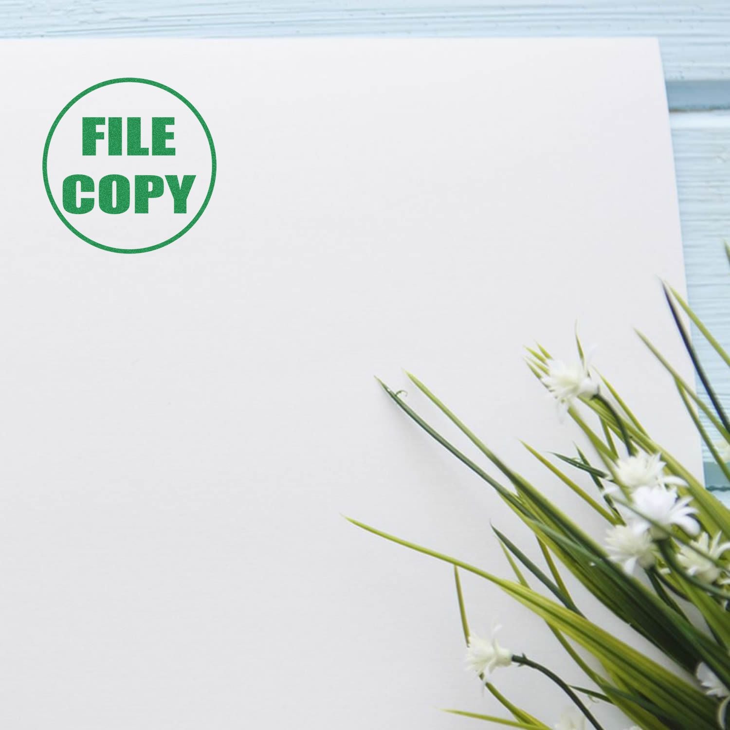 Self Inking Round File Copy Stamp marking FILE COPY in green on a white paper, with green plants in the corner, on a light blue wooden surface.