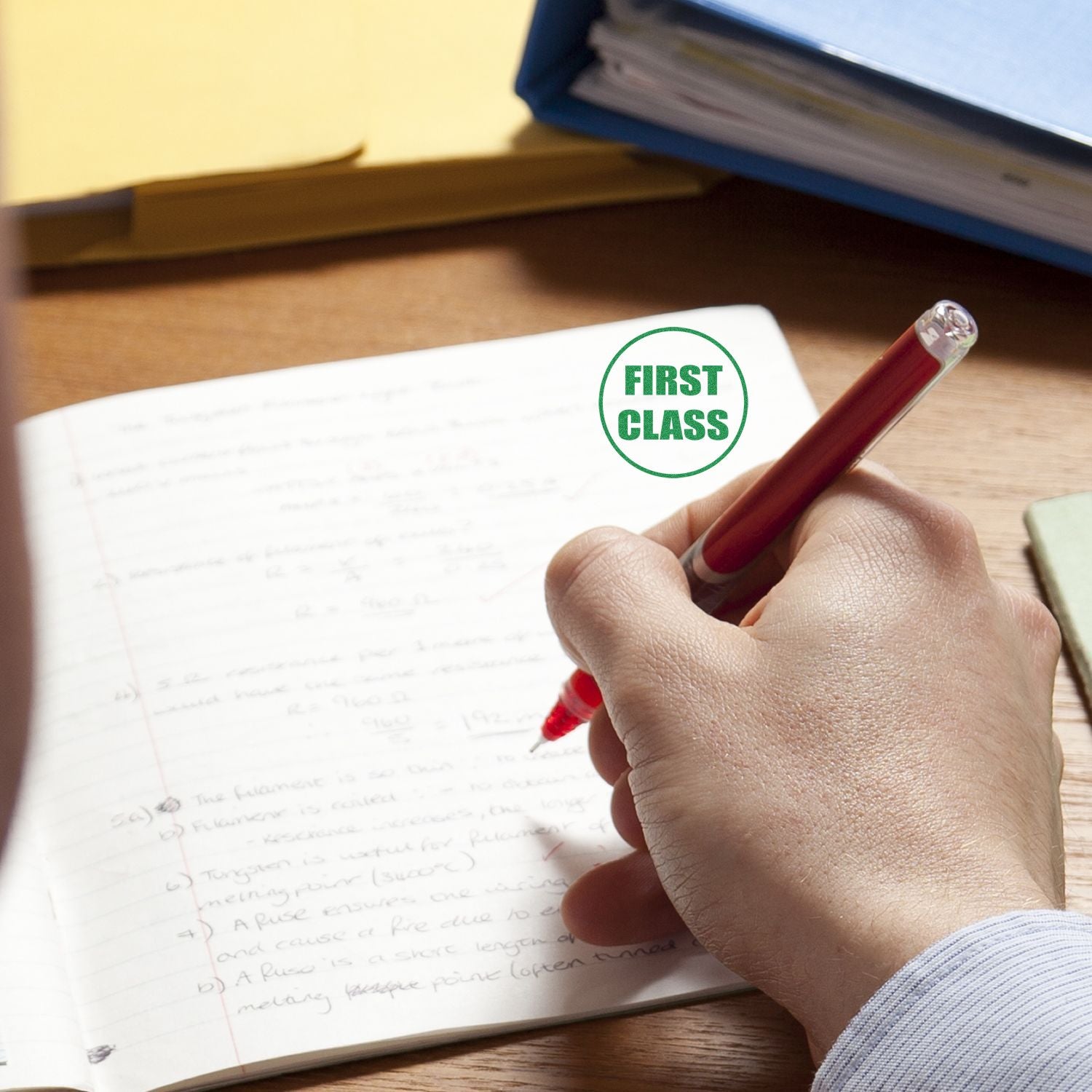 Hand writing in a notebook with a Round First Class Rubber Stamp impression in green ink on the page.