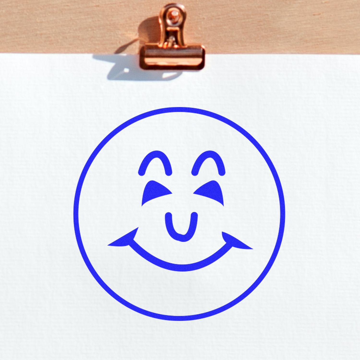 Self Inking Round Happy Face Stamp in blue ink on white paper, clipped to a wooden surface with a copper clip.