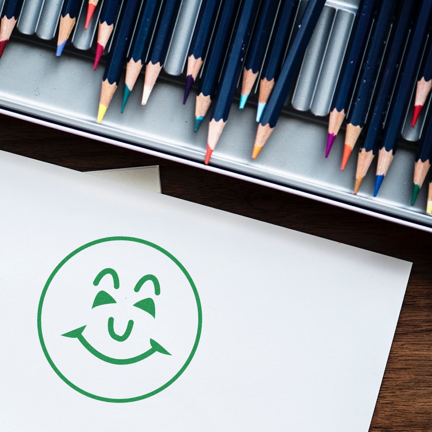 Self Inking Round Happy Face Stamp in green ink on white paper, with colored pencils in the background.