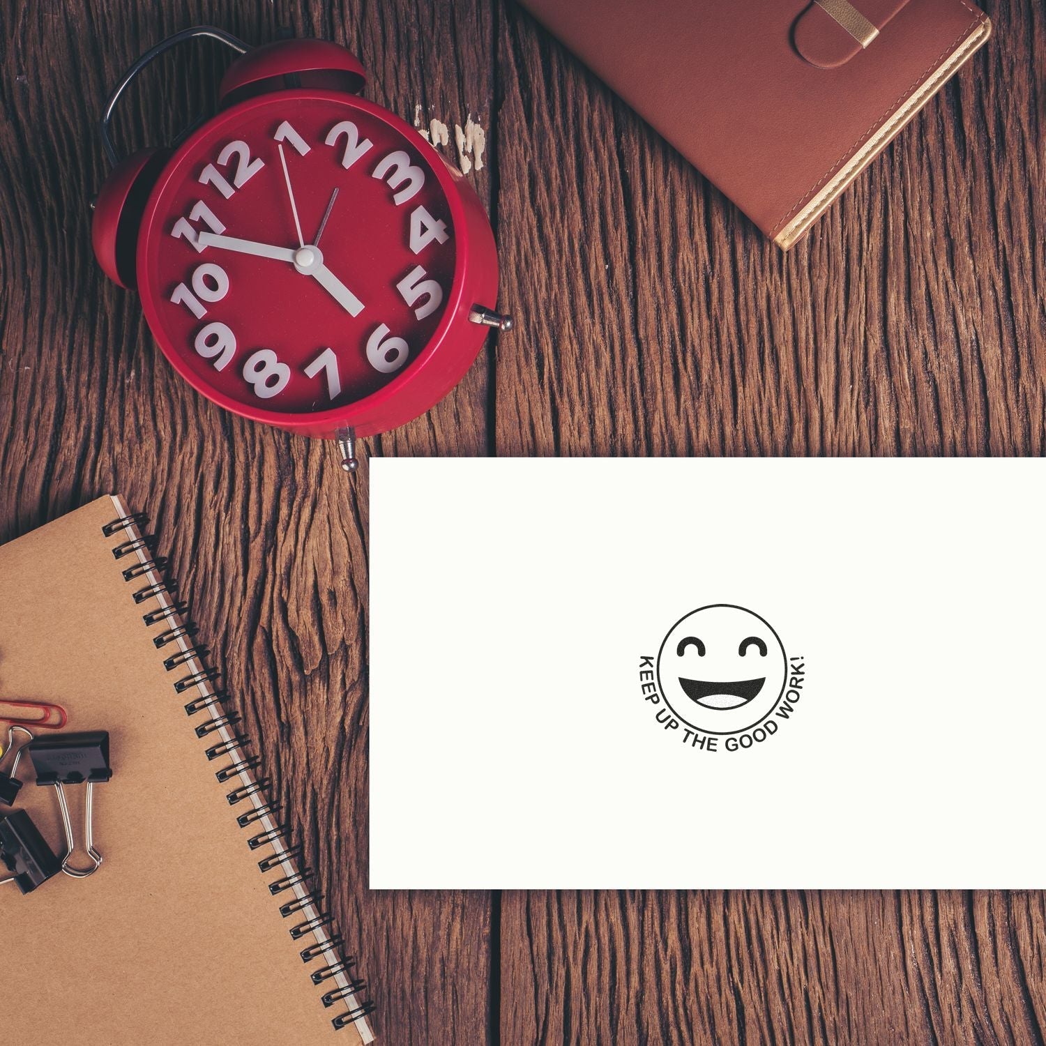 A Self Inking Round Keep up the Good Work Smiley Stamp on white paper, surrounded by a red clock, notebook, and office supplies on a wooden desk.