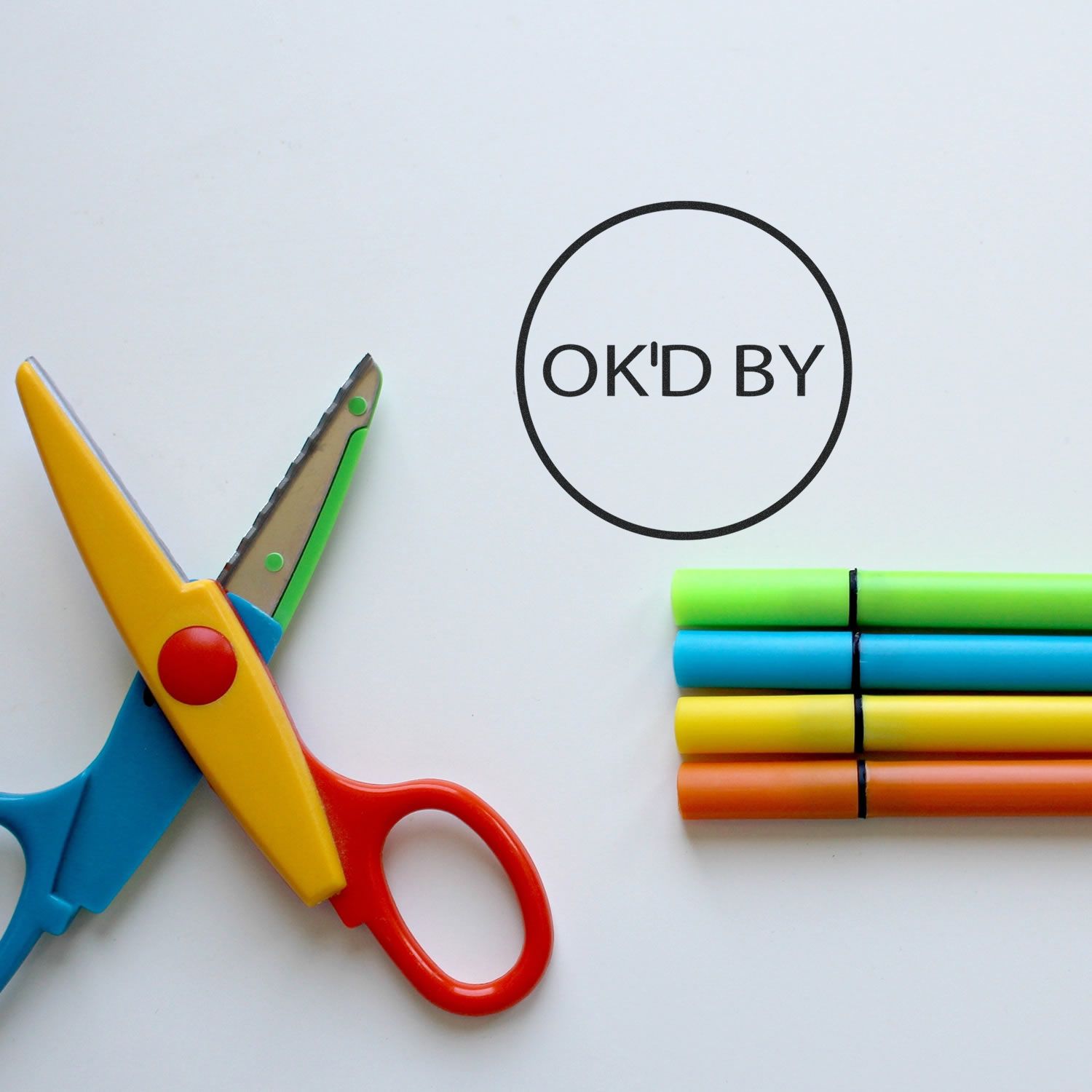 Self Inking Round Ok'd By Stamp on white paper next to colorful scissors and four stacked markers.