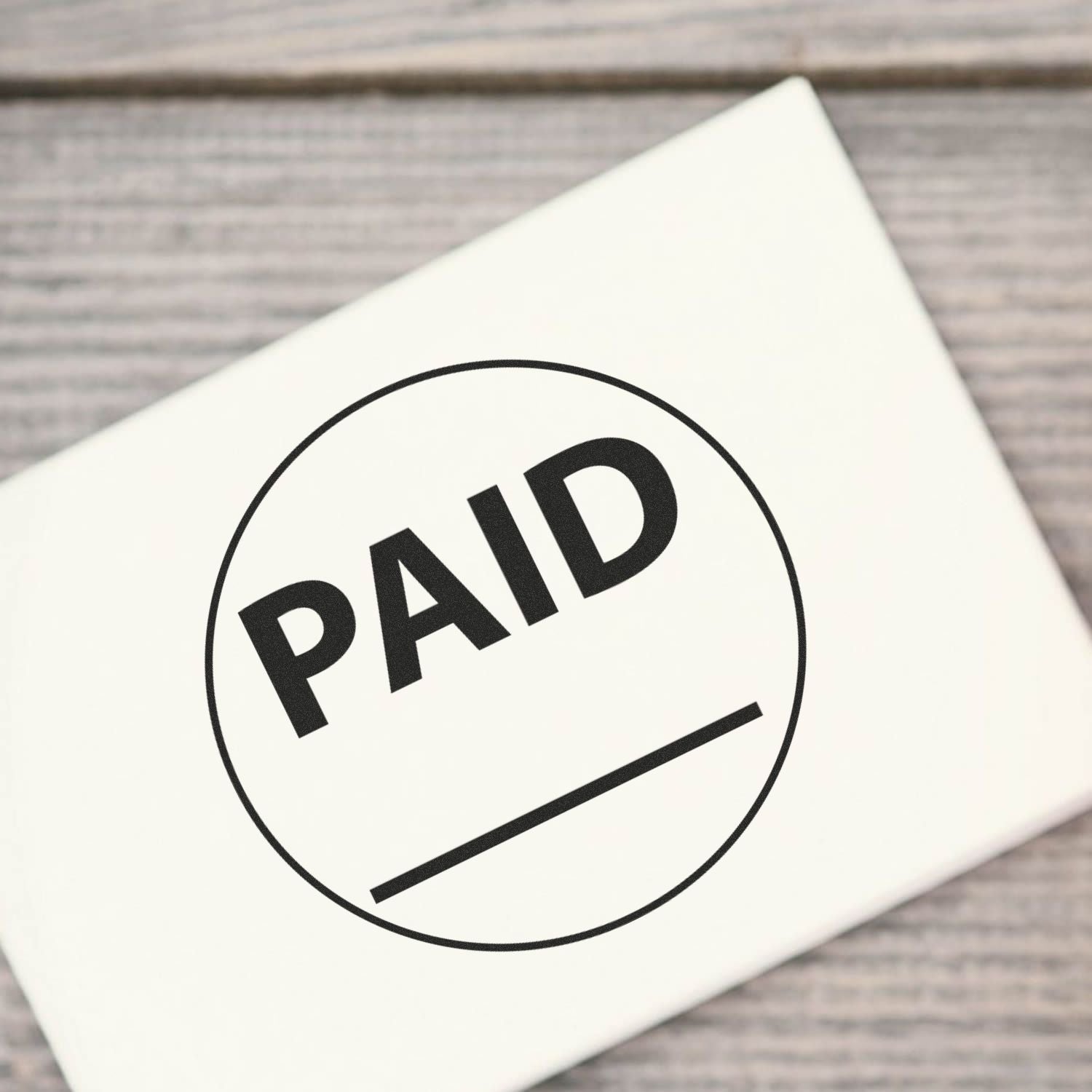 Self Inking Round Paid with Line Stamp marking PAID on a white surface, placed on a wooden background.