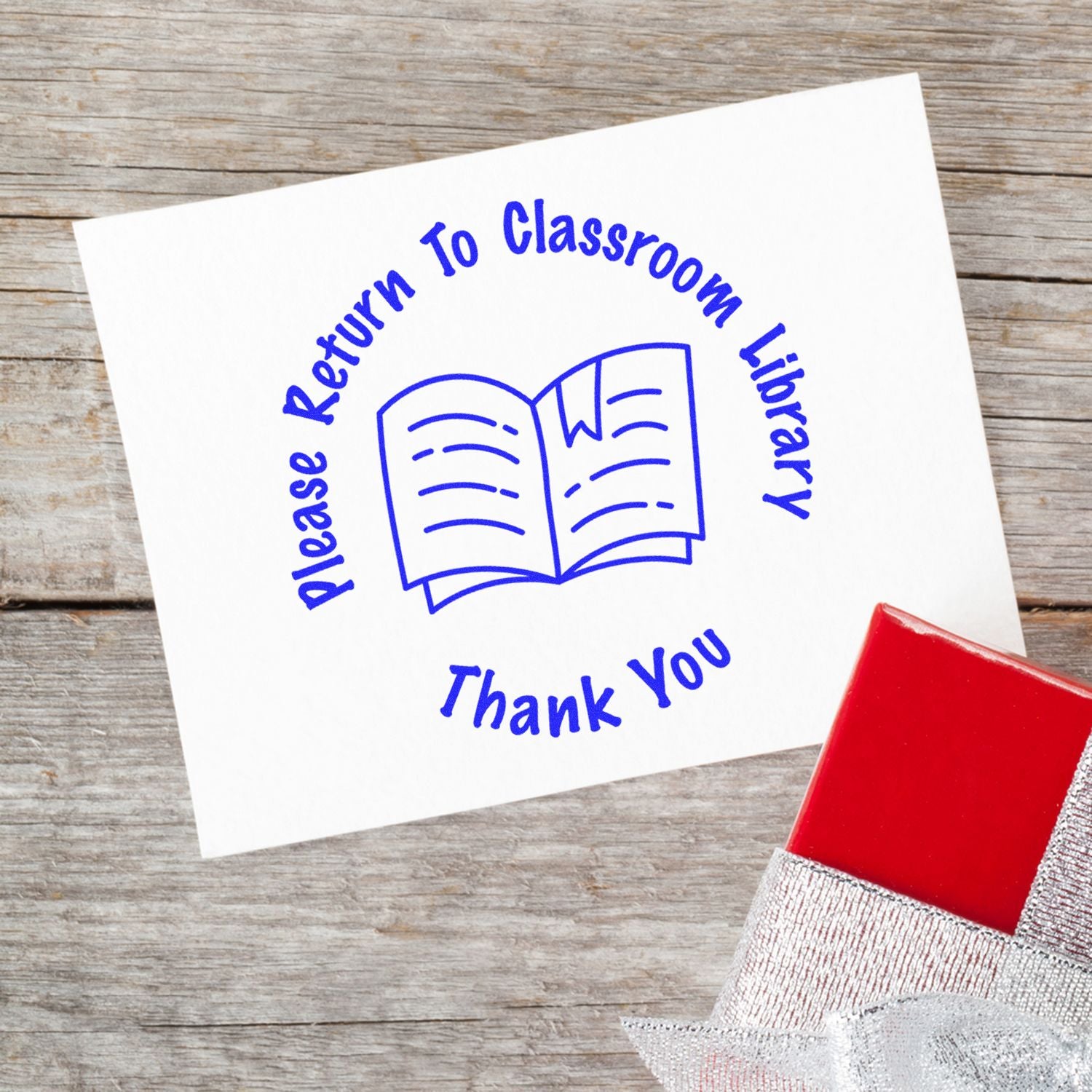 Round Please Return to Classroom Library rubber stamp on a white card with a red ink pad on a wooden surface.