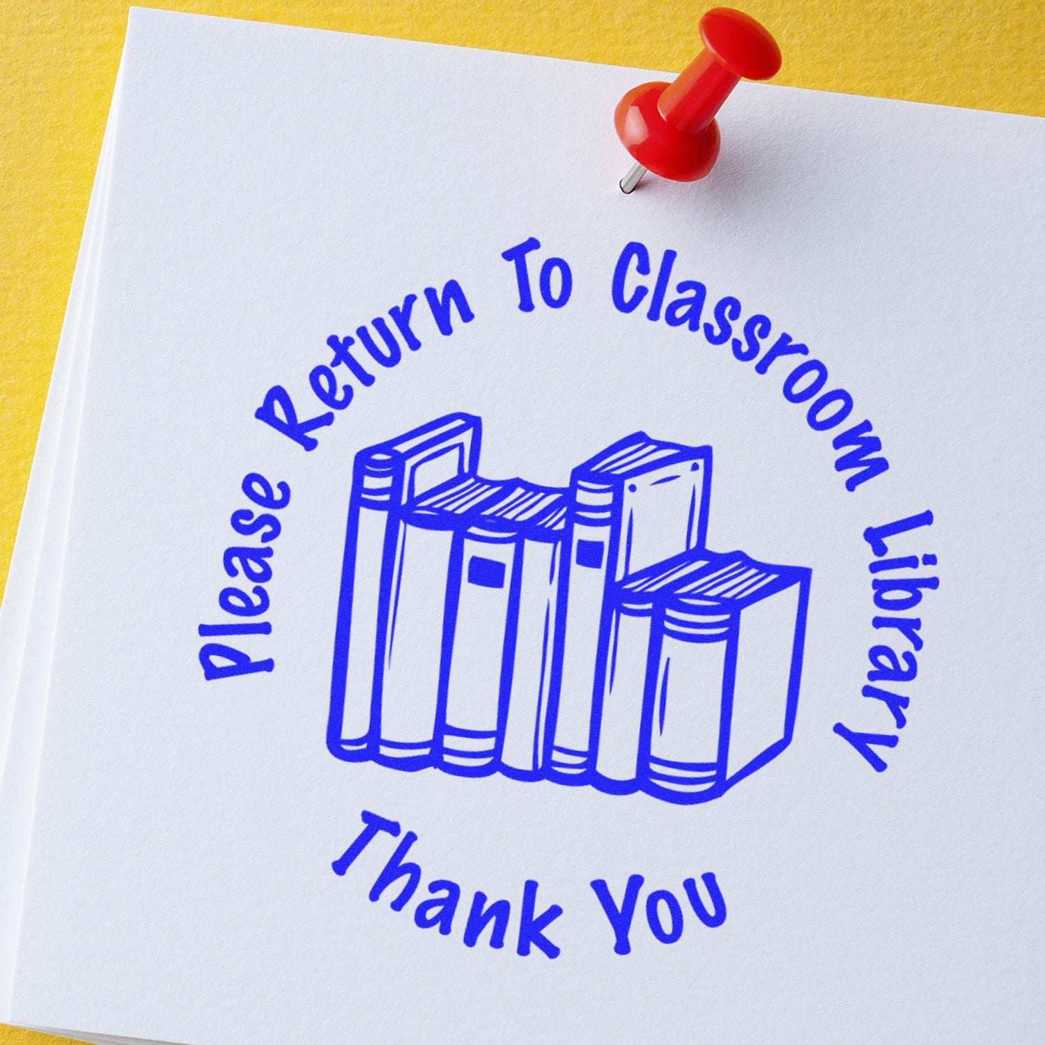 Round Please Return to Classroom rubber stamp with blue ink on white paper, featuring a stack of books and a red pushpin.