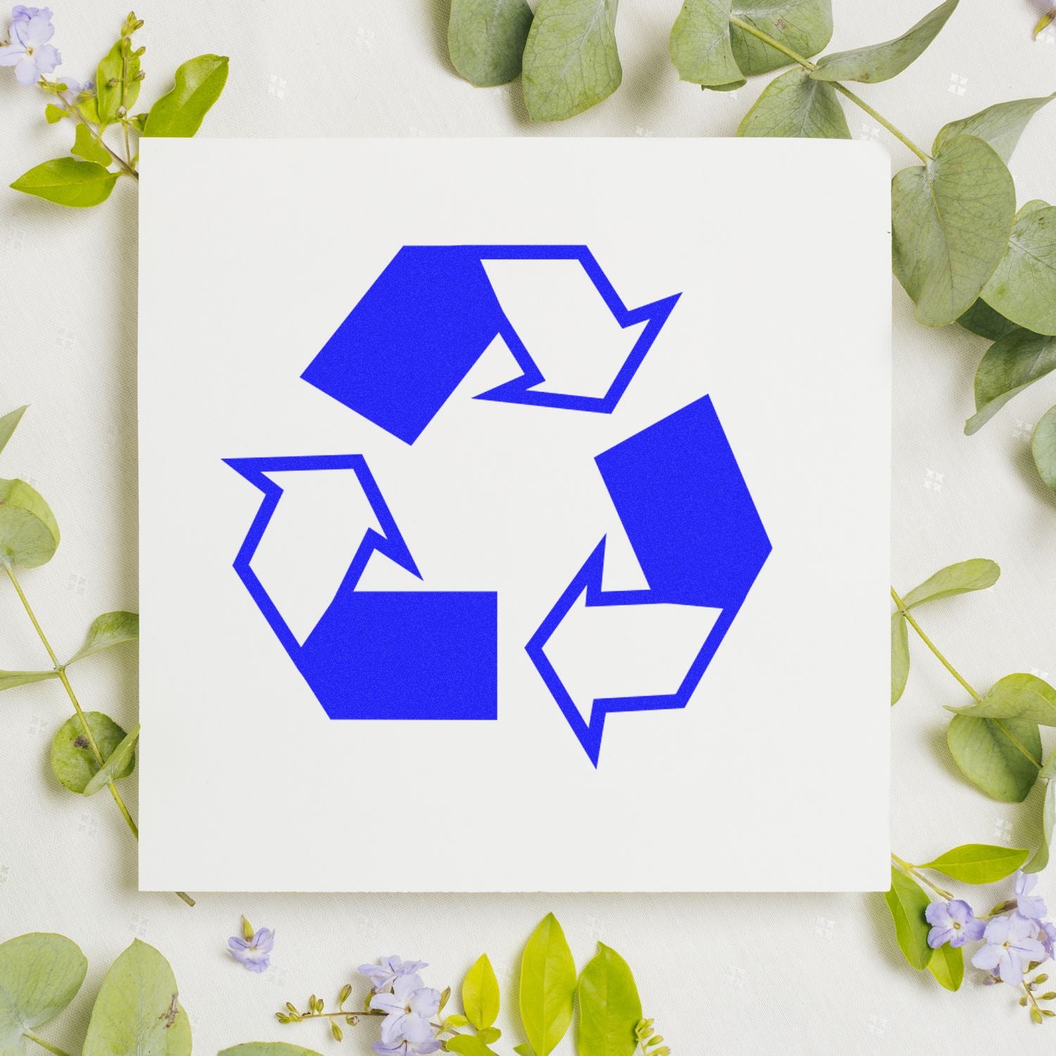 Round Recycle Rubber Stamp with a blue recycling symbol on white paper, surrounded by green leaves and small purple flowers.