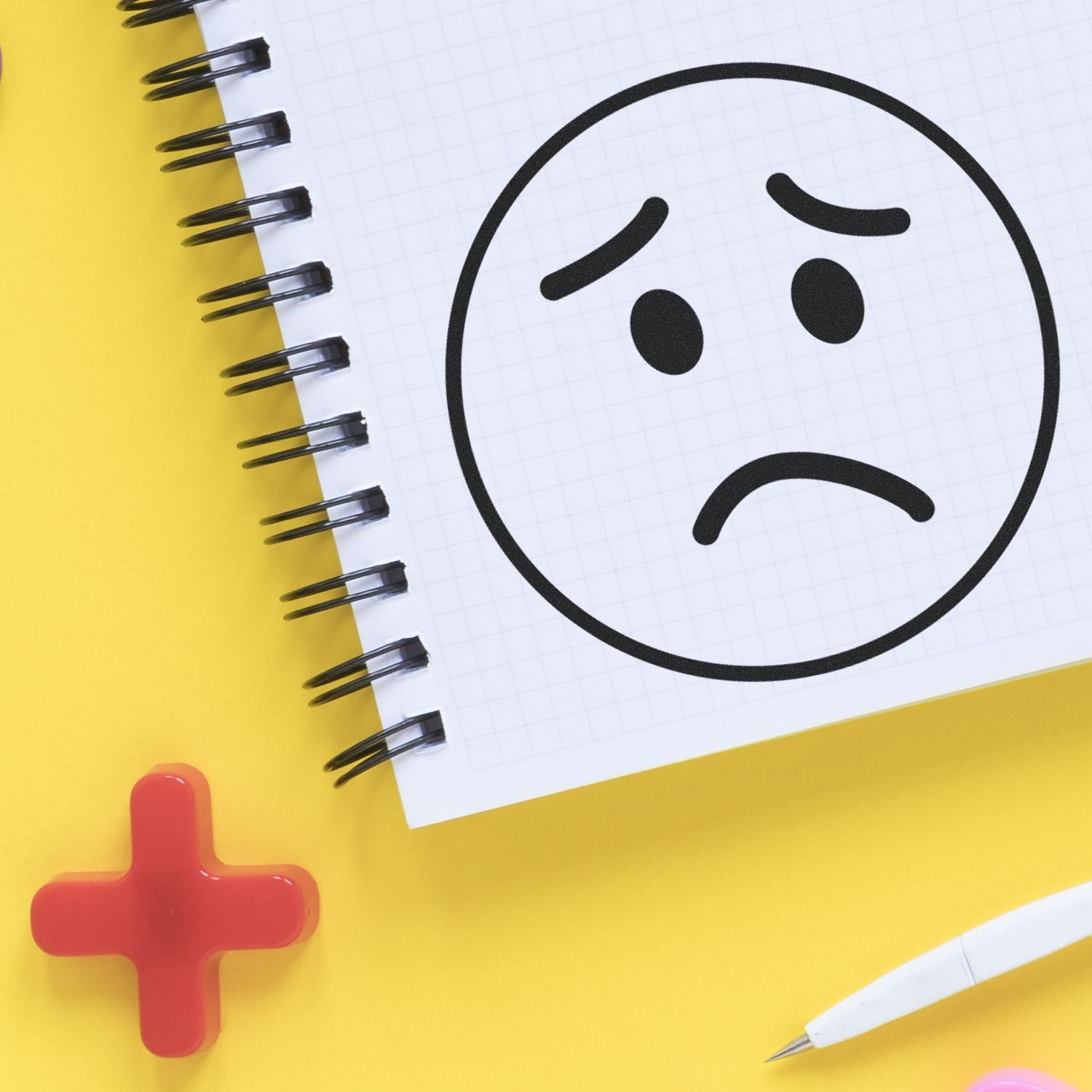 A notebook with a stamped sad face using the Self Inking Round Sad Face Stamp, placed on a yellow surface with a red cross and white pen nearby.