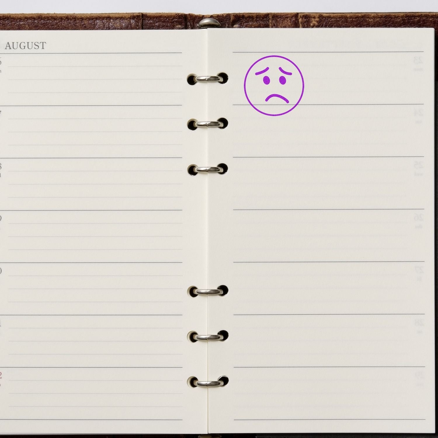 Self Inking Round Sad Face Stamp in purple ink on a blank page of a planner, highlighting a specific date in August.