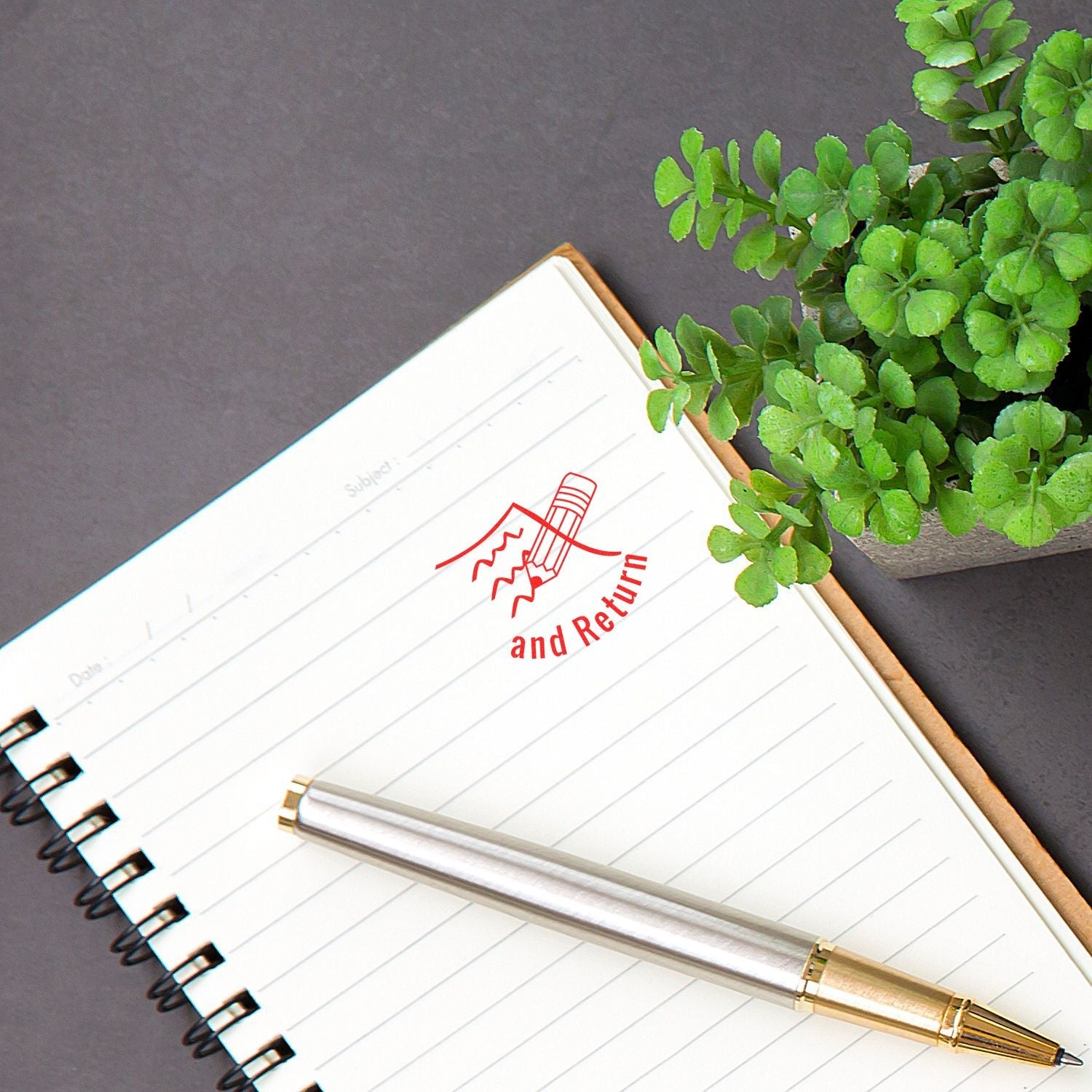 A notebook with a Sign and Return mark from the Self Inking Round Curious Smiley Stamp, next to a pen and a small potted plant.