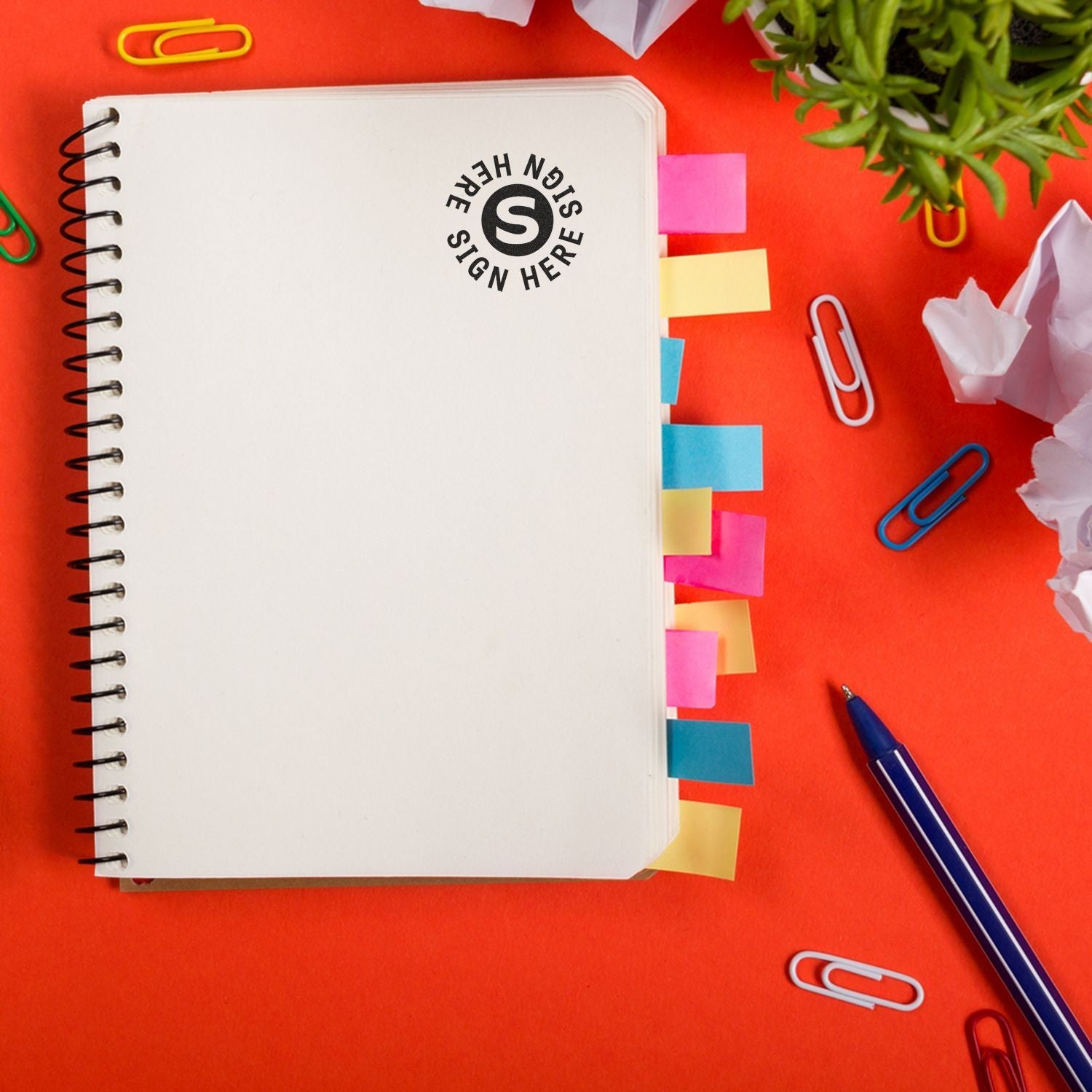 Self Inking Round Sign Here Sign Here Stamp on an open spiral notebook with colorful sticky tabs, surrounded by paper clips and a pen.