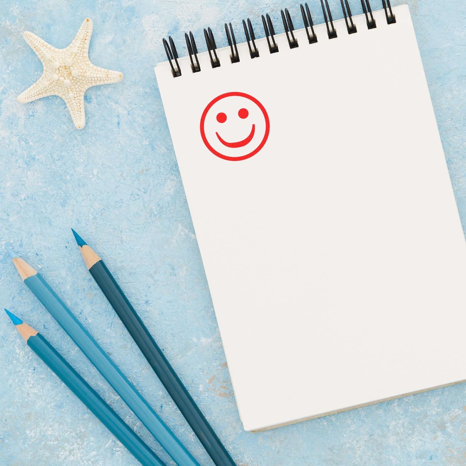 A notebook with a red smiley face stamped using the Self Inking Round Smiley Stamp, placed next to blue pencils and a starfish on a blue background.