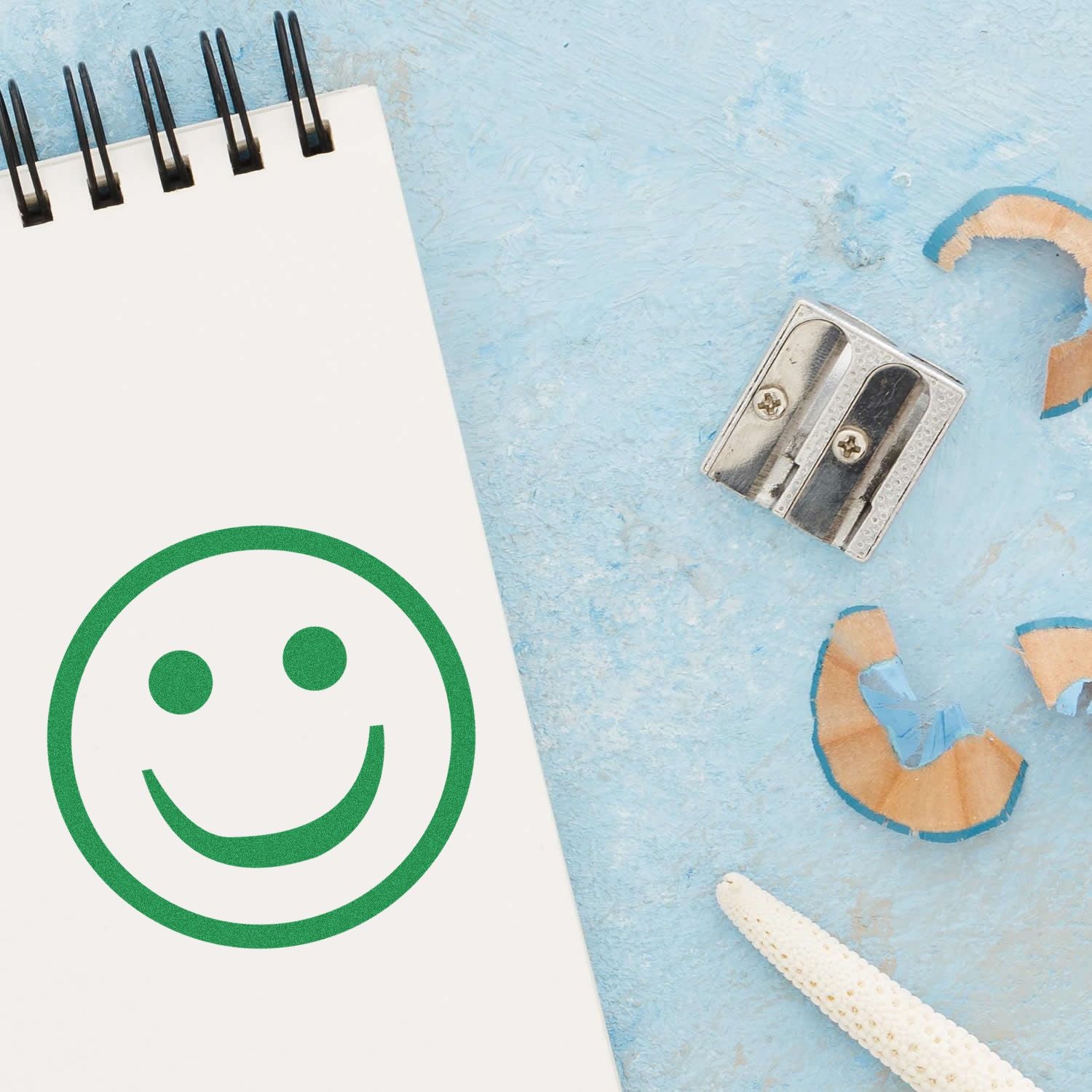 Round Smiley Rubber Stamp in green ink on a notebook page, next to a pencil sharpener and shavings on a light blue surface.