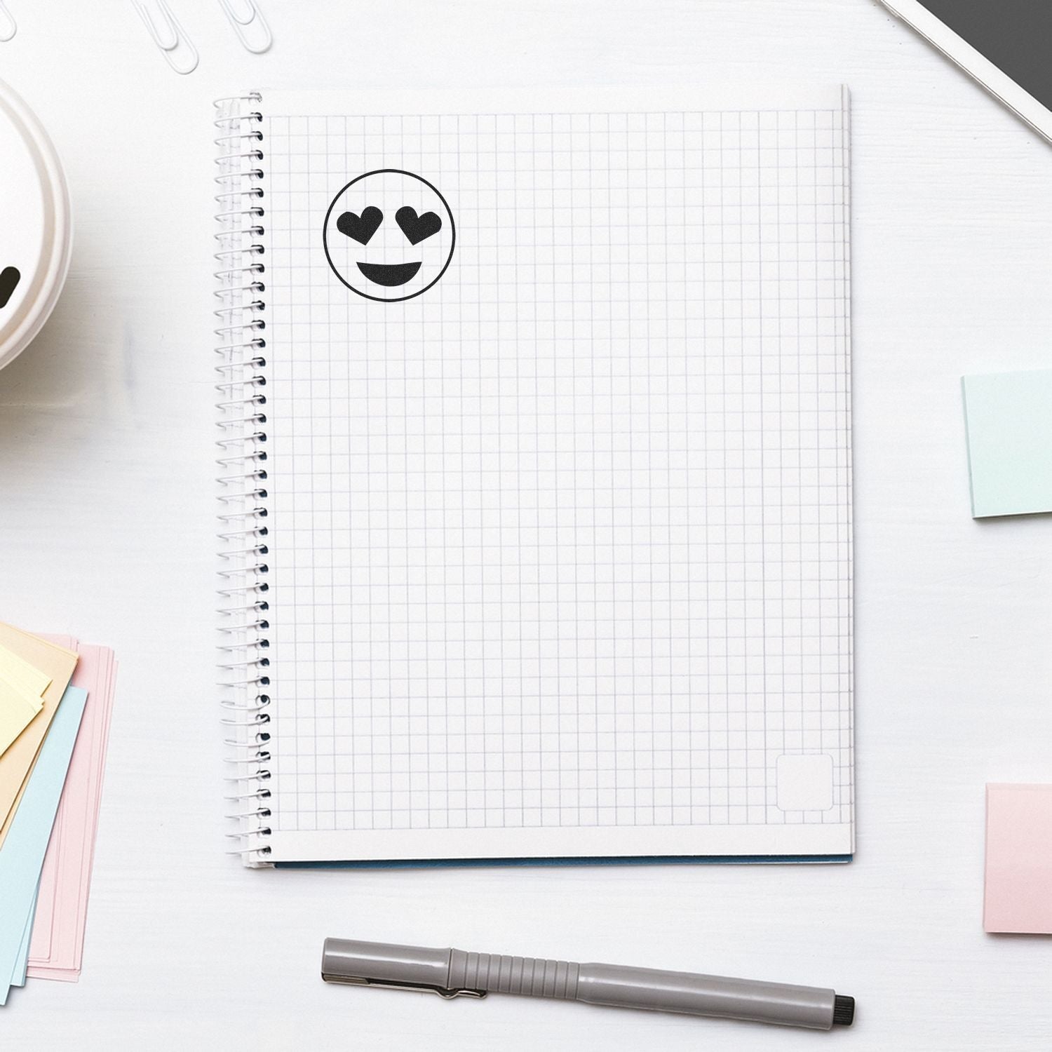 Self Inking Round Smiley with Hearts Stamp used on a graph paper notebook, surrounded by a pen, sticky notes, and a smartphone.