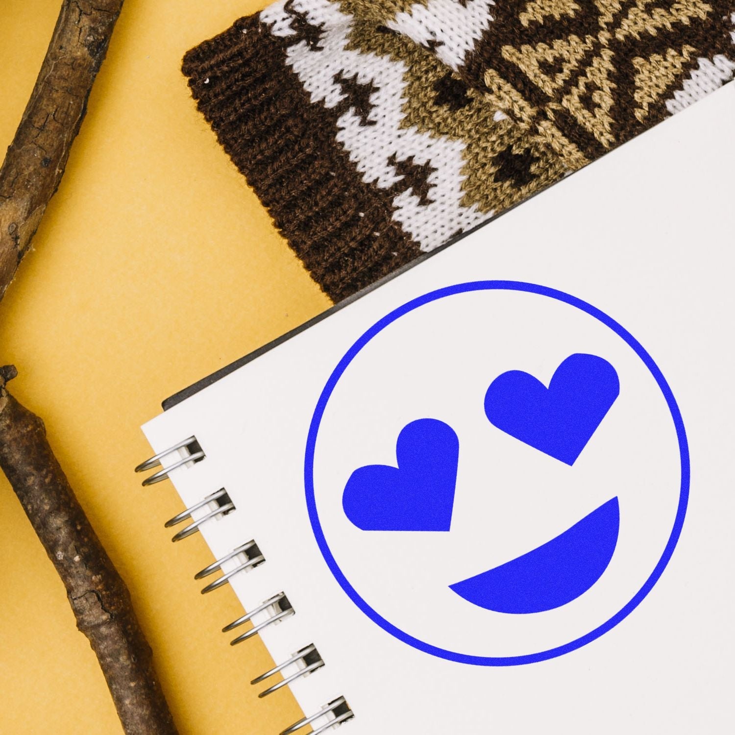 Self Inking Round Smiley with Hearts Stamp in blue ink on a white notebook, next to a knitted sleeve and wooden sticks.