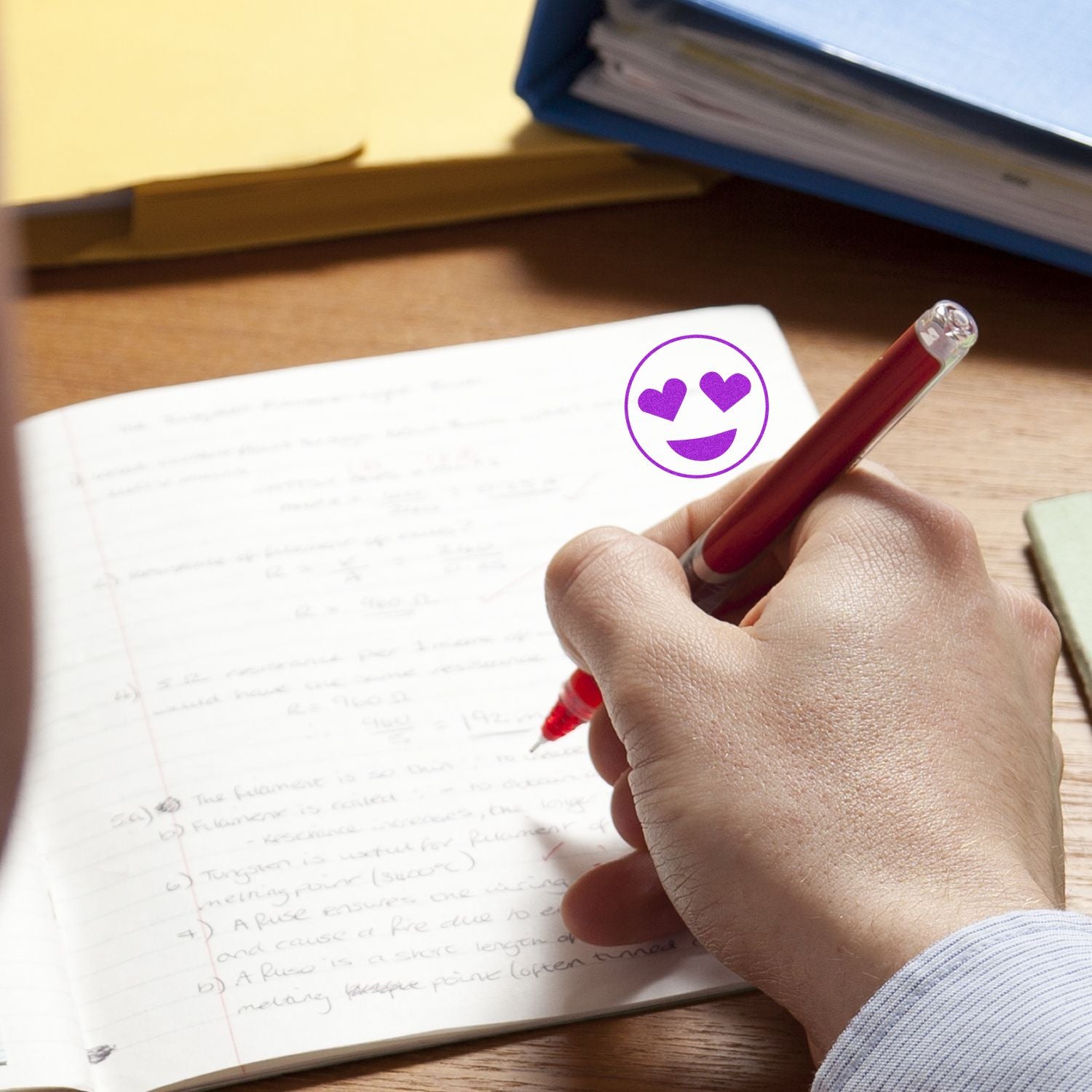 A hand writing in a notebook with a red pen, next to a purple Round Smiley with Hearts Rubber Stamp impression on the page.