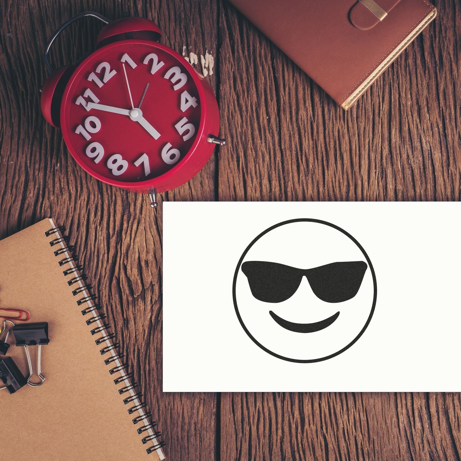 Self Inking Round Smiley with Sunglasses Stamp on white paper, placed on a wooden desk with a red clock, notebook, and office supplies.