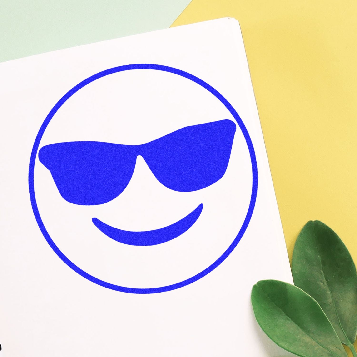 Self Inking Round Smiley with Sunglasses Stamp in blue ink on white paper, with green leaves and yellow background.