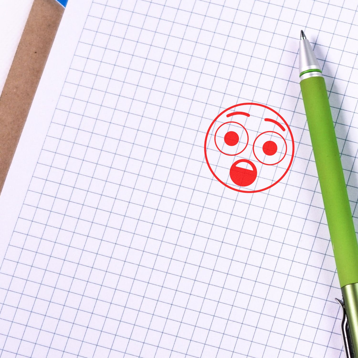 A green pen lies on graph paper stamped with a red Round Surprised Smiley Rubber Stamp, showing a surprised face.