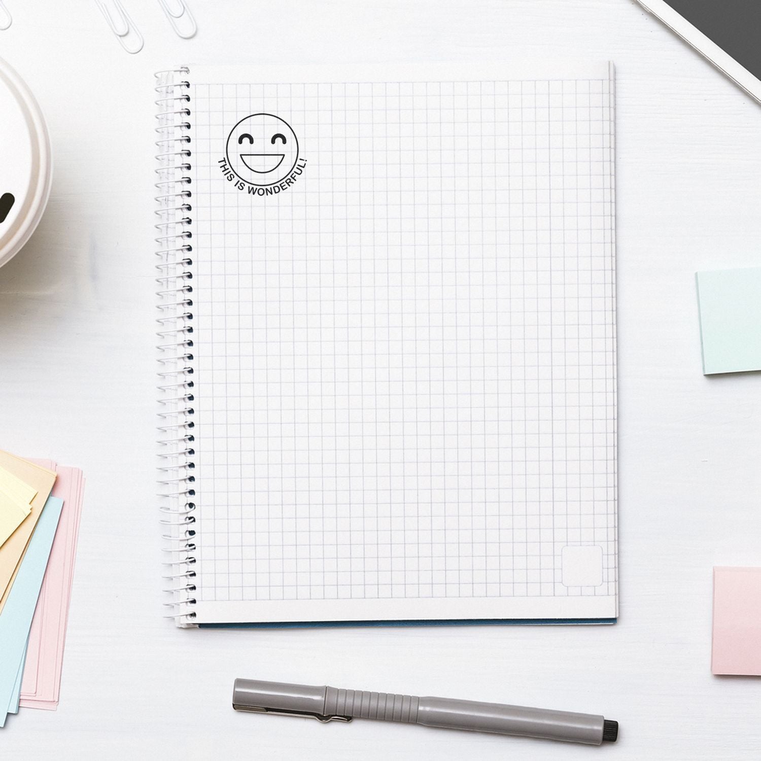 Round This is Wonderful Smiley Self-Inking Stamp on a grid notebook page, surrounded by sticky notes, a pen, and a smartphone.