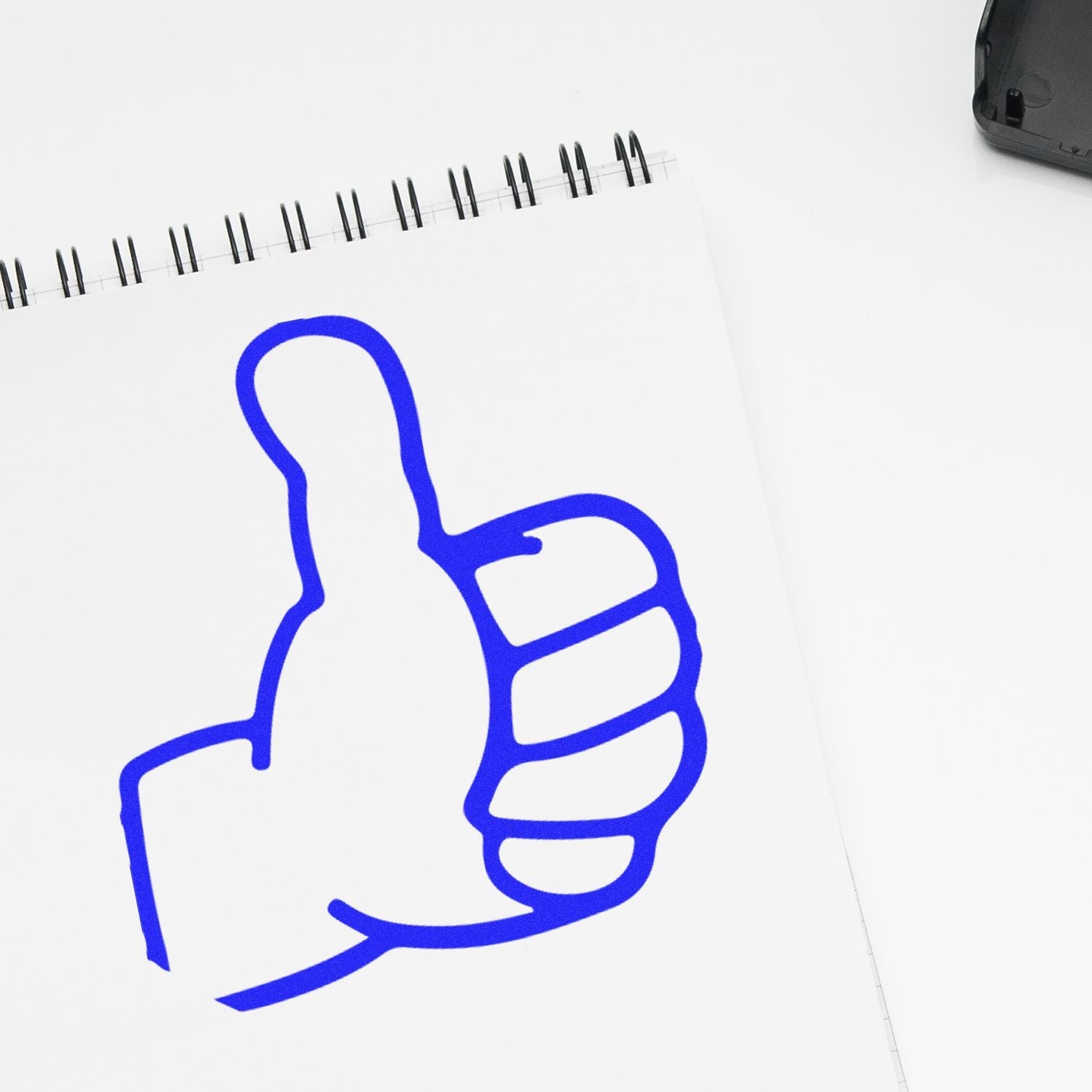 A blue thumbs-up mark on white paper made with the Self Inking Round Thumbs Up Stamp, next to a black stamp case.