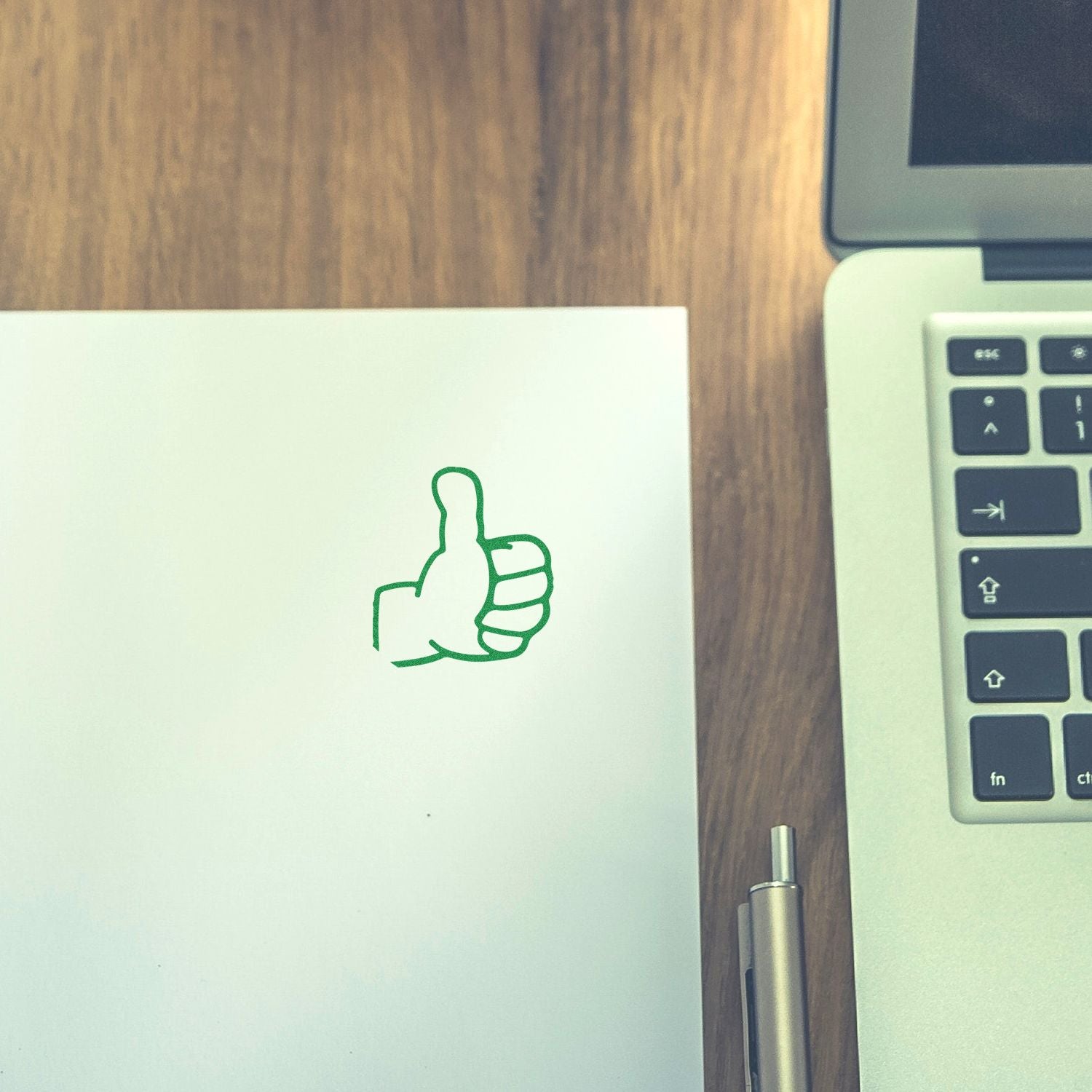 Round Thumbs Up Rubber Stamp in green ink on white paper, next to a silver pen and a laptop on a wooden desk.