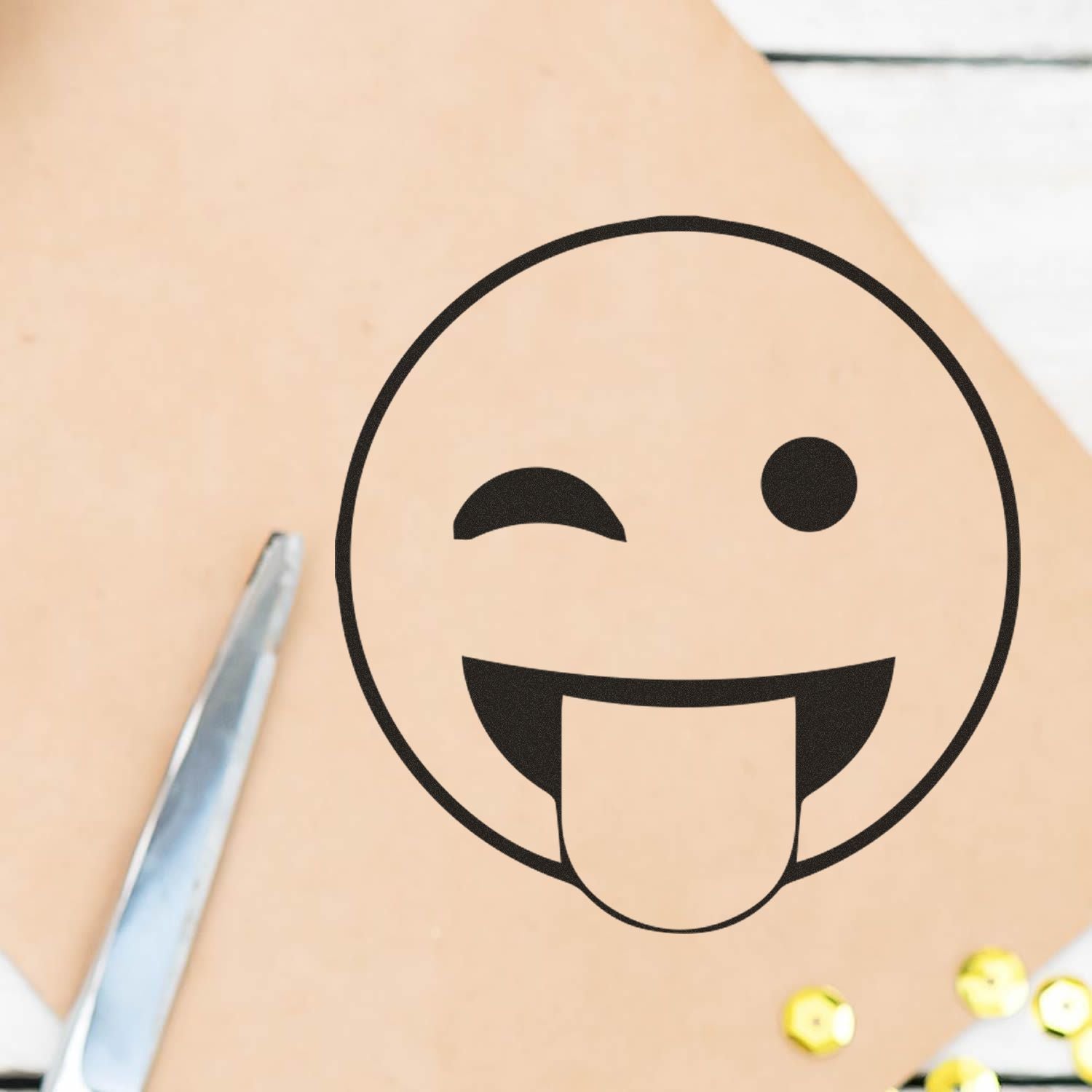 Round Tongue Out Smiley Rubber Stamp on brown paper, next to a pair of scissors and yellow sequins.