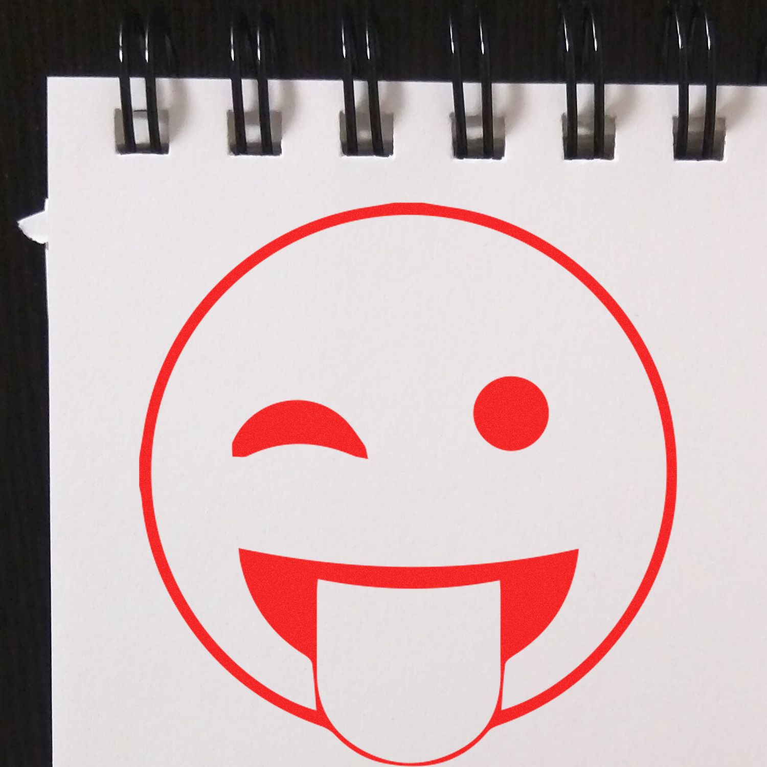 Self Inking Round Tongue out smiley Stamp in red ink on white notepad with black spiral binding.