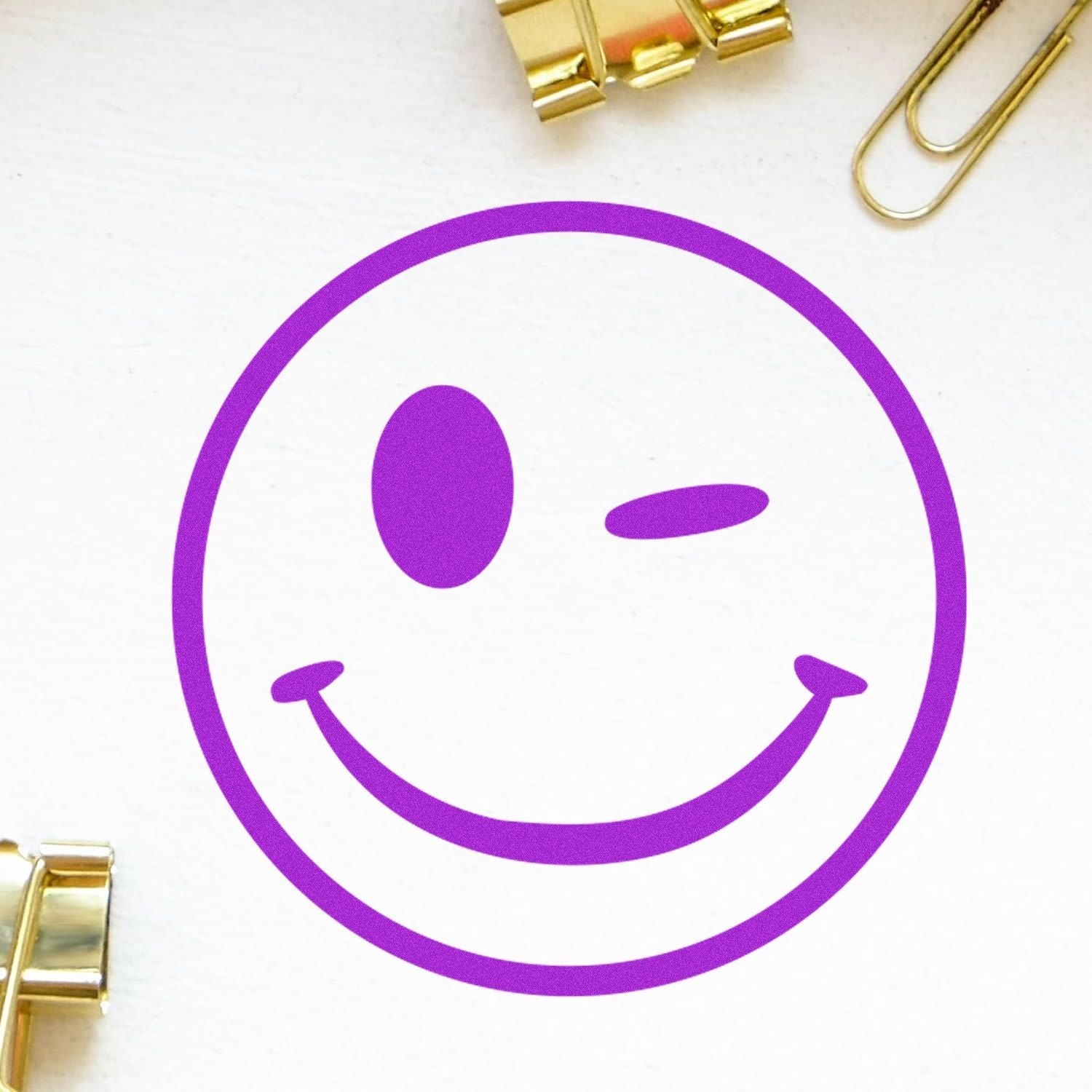 A purple winking smiley face stamped on white paper, created with the Self Inking Round Winking Smiley Stamp, surrounded by gold paper clips.