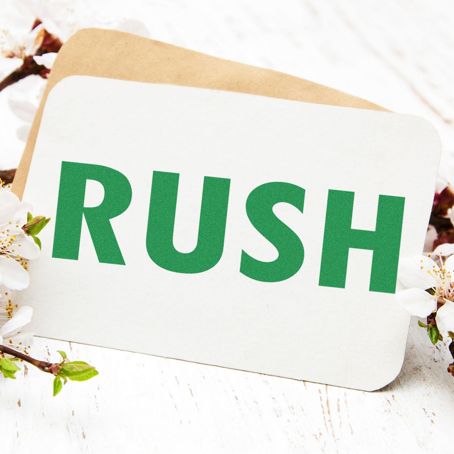 Large Pre-Inked Rush Stamp in green ink on a white card, surrounded by white flowers and a brown envelope on a wooden surface.